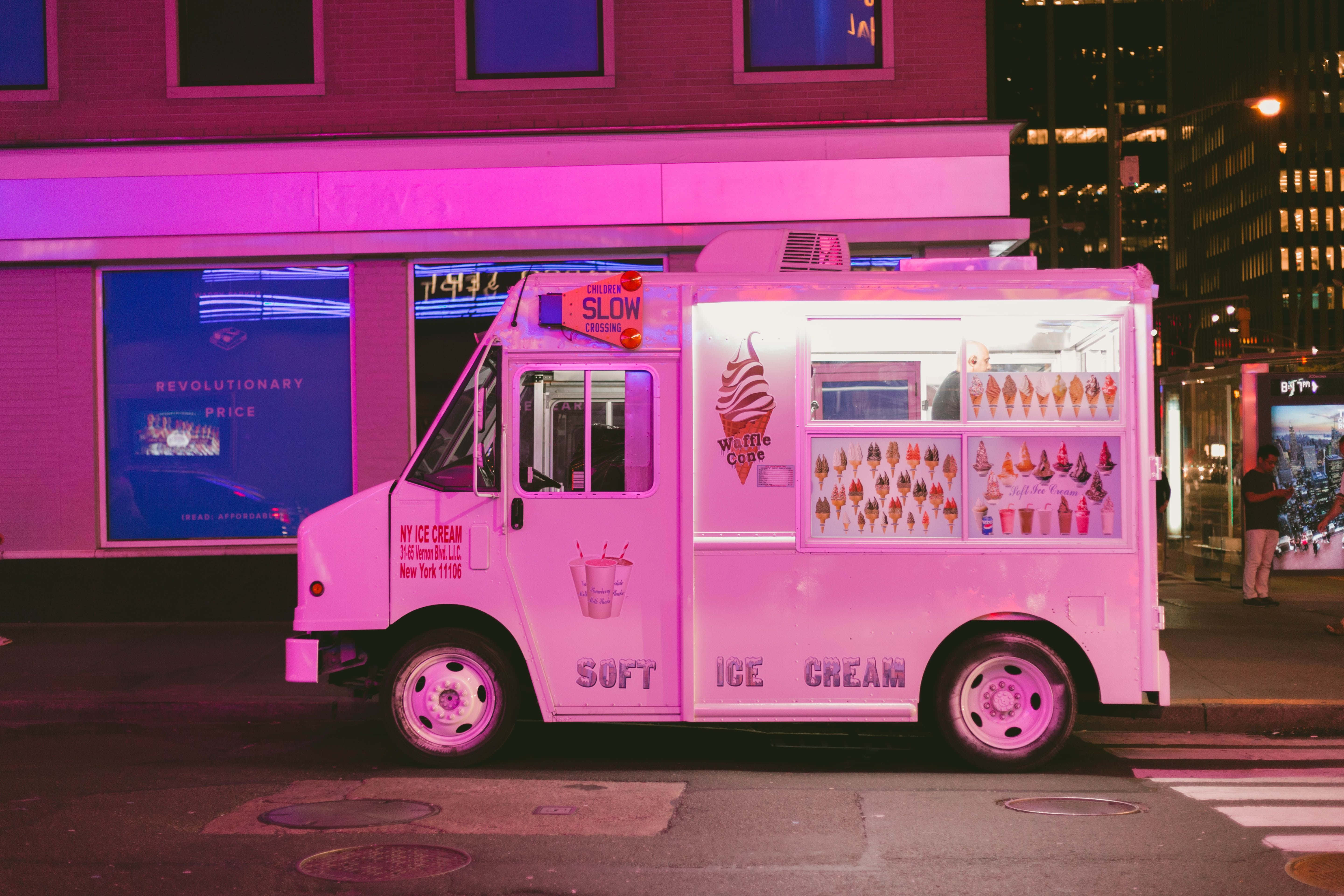 Ice Cream Truck 桌布 壁紙 — 一輛鮮豔的粉紅色冰淇淋車停在夜晚霓虹燈閃爍的城市街道上，車窗發光，背景是城市建築。