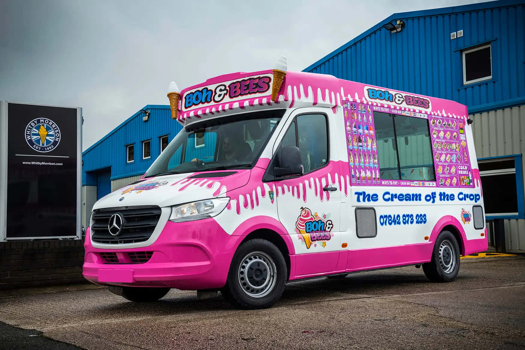 Ice Cream Truck 桌布 壁紙 — 一輛鮮豔的粉紅色和白色冰淇淋車，印有 'Boh & Bees' 標誌，停放在藍色工業建築外，天空多雲。