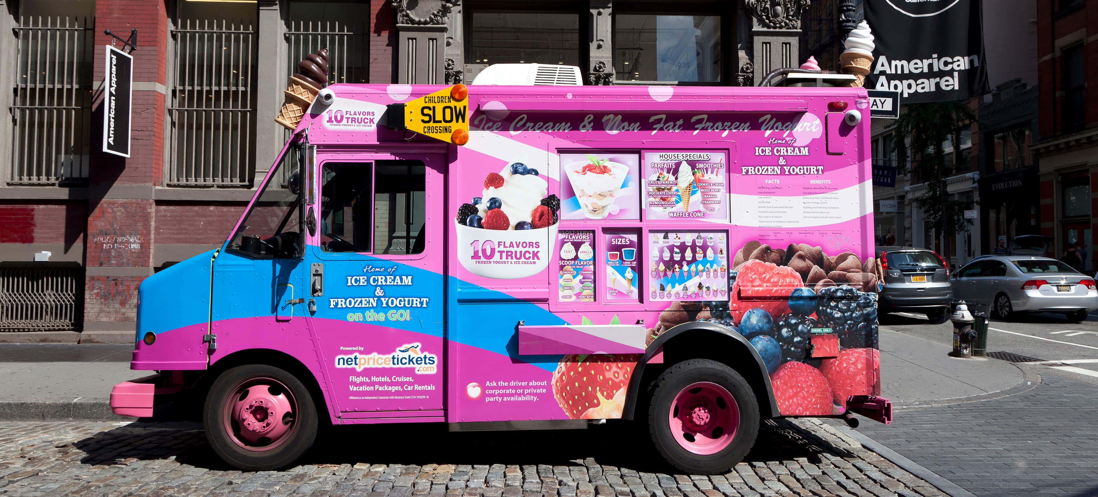 Ice Cream Truck 桌布 壁紙 — 一輛鮮豔的粉紅色和藍色冰淇淋車停在鵝卵石城市街道上，帶有色彩繽紛的甜品圖案和「SLOW」標誌。