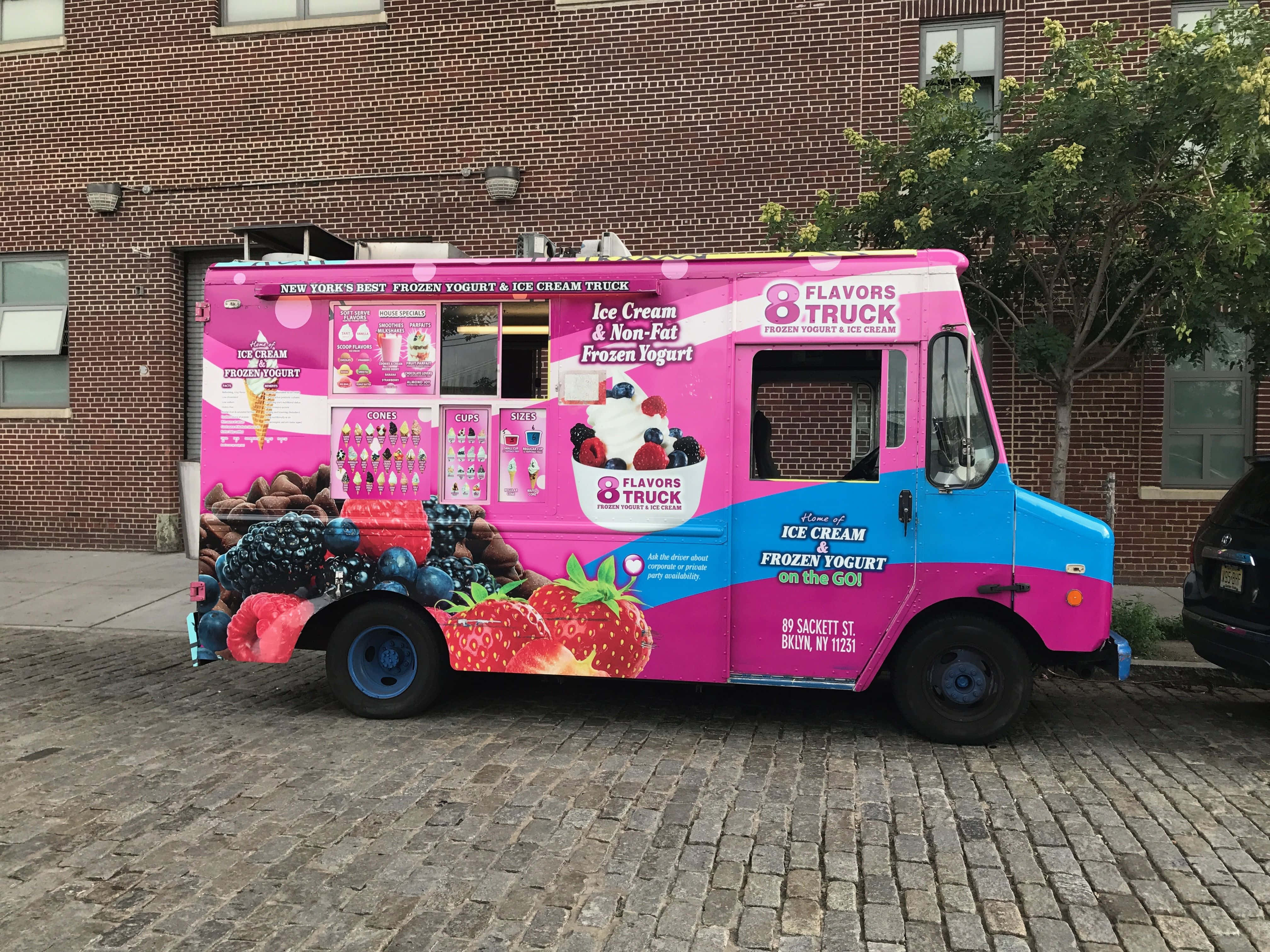 Ice Cream Truck 桌布 壁紙 — 一輛鮮豔的粉紅色和藍色冰淇淋車停泊在鵝卵石街道上，旁邊是一棟帶有水果圖案和菜單顯示屏的磚砌建築。