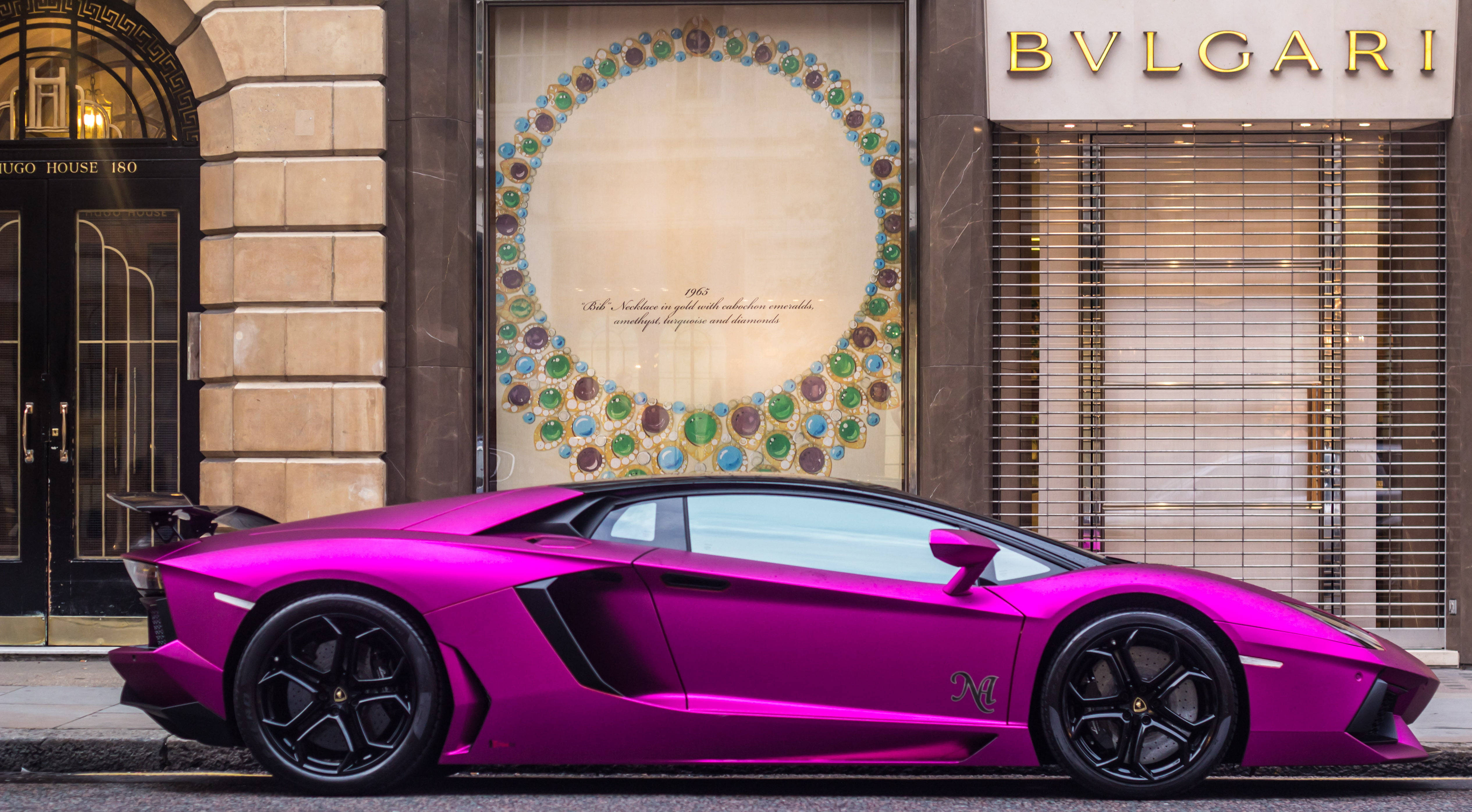 Bvlgari 桌布 壁紙 — 一輛鮮豔的紫紅色藍寶堅尼 Aventador 停放在城市街道上，前方是 Bulgari 珠寶店，店內有一個大型圓形寶石展示。