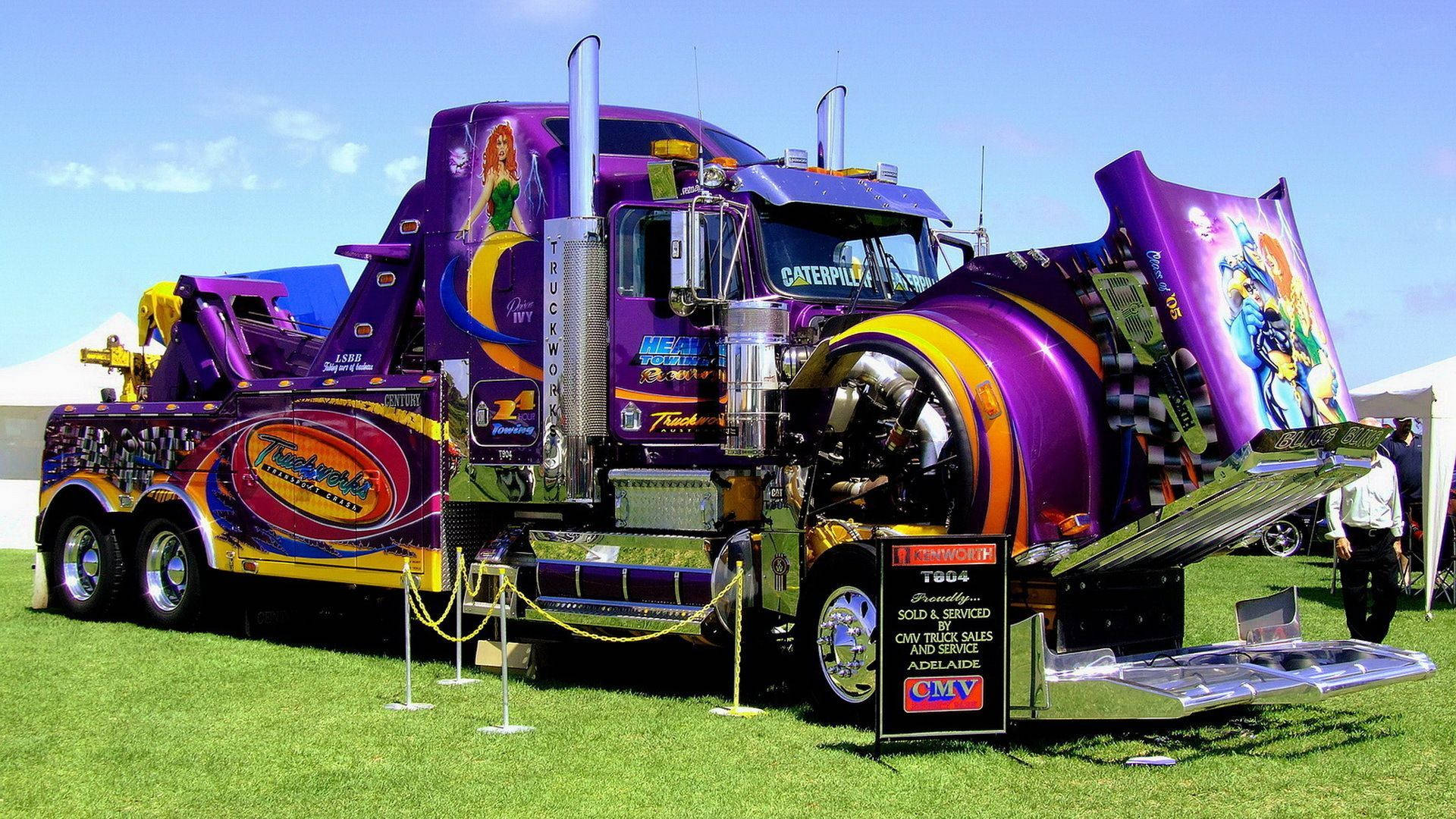 Kenworth 桌布 壁紙 — 一輛鮮豔的紫色和金色定制拖車，車廂上有卡通藝術，在晴朗藍天下的綠色草地上戶外展覽中展示。