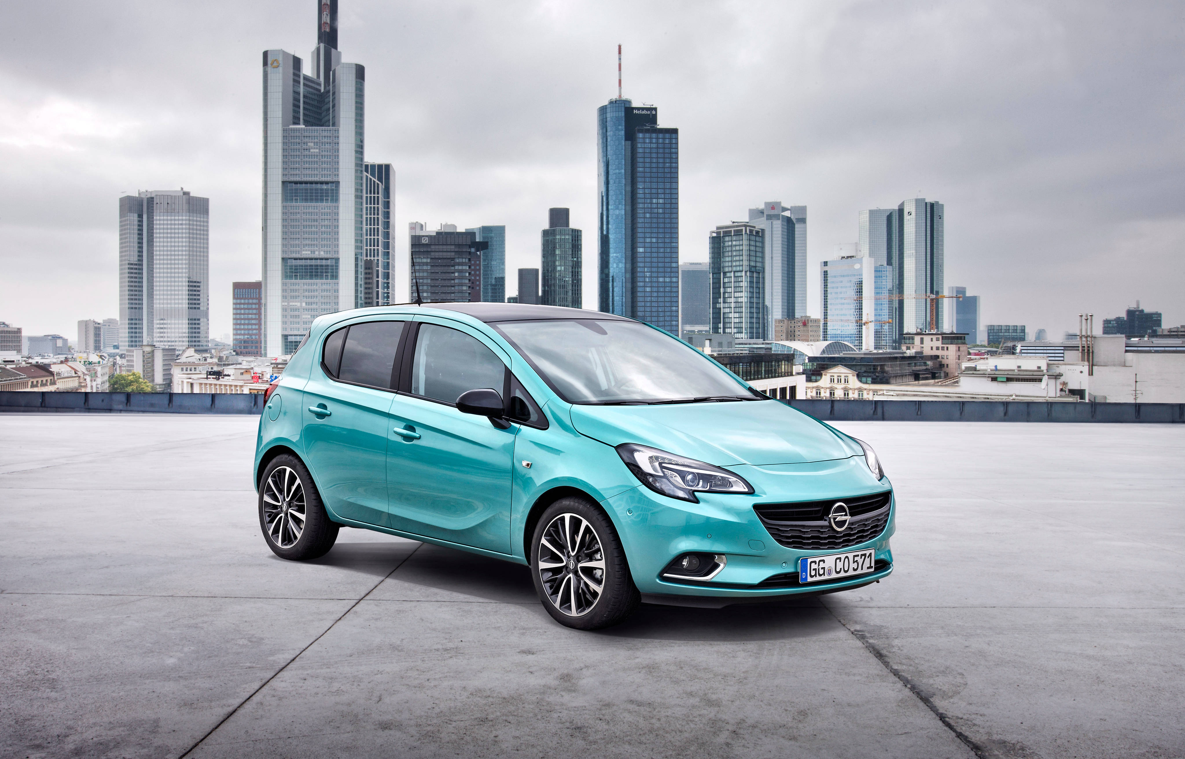 Opel 桌布 壁紙 — 一輛鮮豔的藍綠色歐寶 Corsa 停放在屋頂上，法蘭克福的現代摩天大樓在陰天籠罩下若隱若現。
