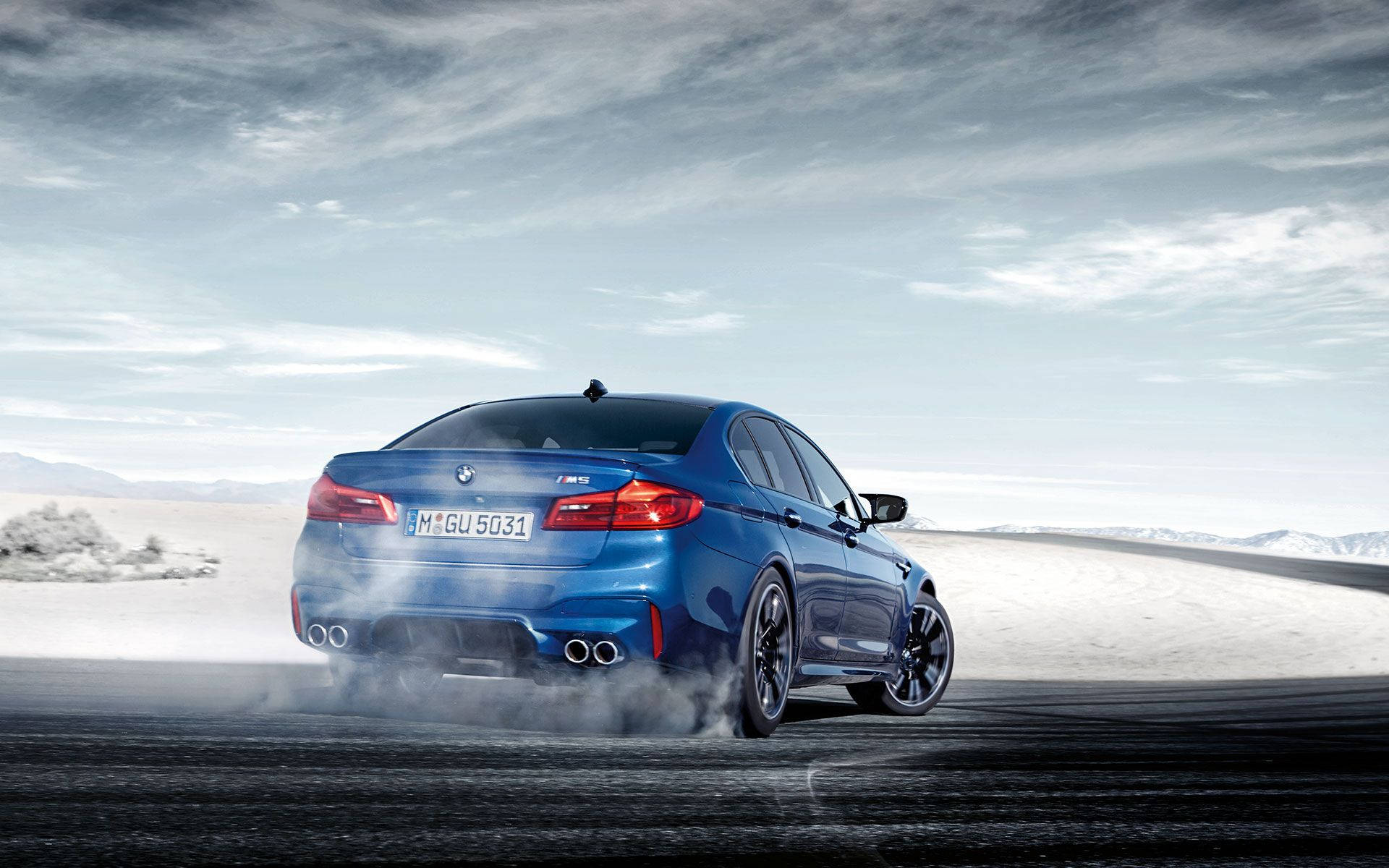 m 桌布 壁紙 — 一輛鮮豔的藍色BMW M5在陰天下的黑暗賽道上漂移，輪胎冒出滾滾濃煙，背景是廣闊的雪沙漠。