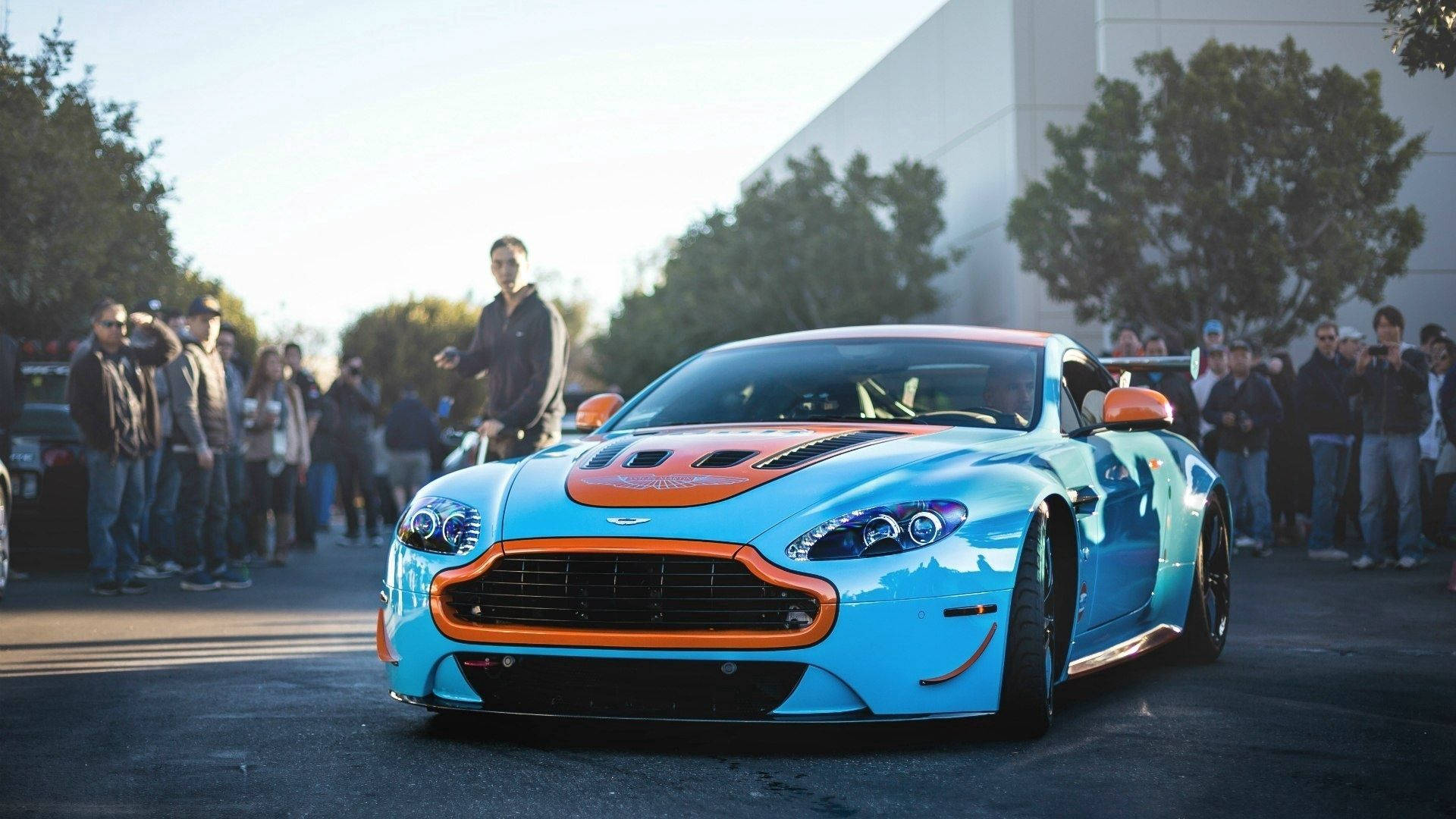 奧斯頓馬丁 桌布 壁紙 — 一輛鮮豔的藍色和橙色Aston Martin跑車在金色陽光下閃閃發光，圍觀者聚集在鋪砌的場地上。