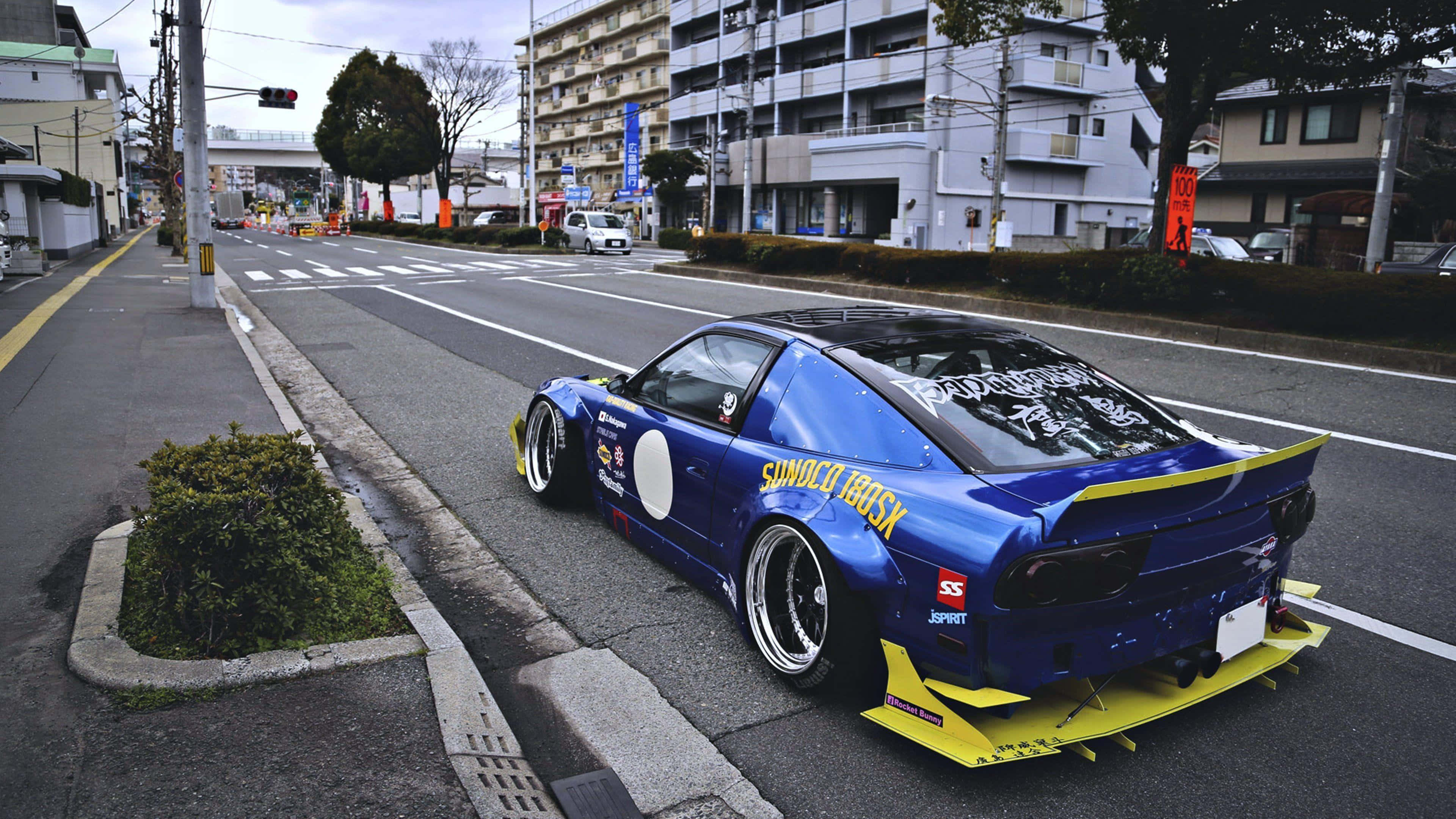Nissan 180sx 桌布 壁紙 — 一輛鮮豔的藍黃色改裝跑車停泊在城市街道上，背景有建築物和樹木。