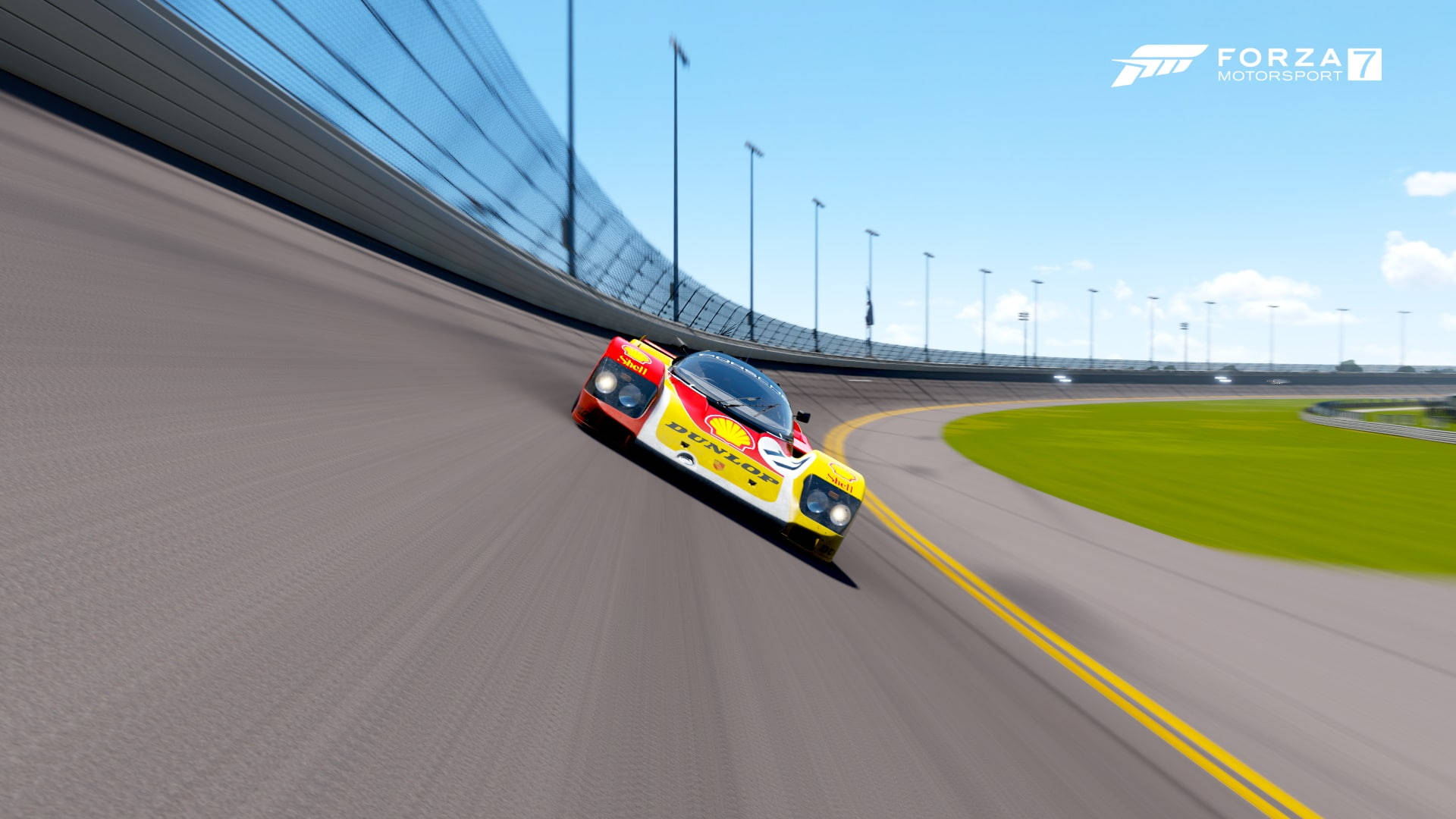 Motorsport 桌布 壁紙 — 一輛鮮豔的黃色和紅色賽車在晴朗的藍天下，以綠色草地和體育場燈光為背景，在彎曲的瀝青賽道上飛馳。