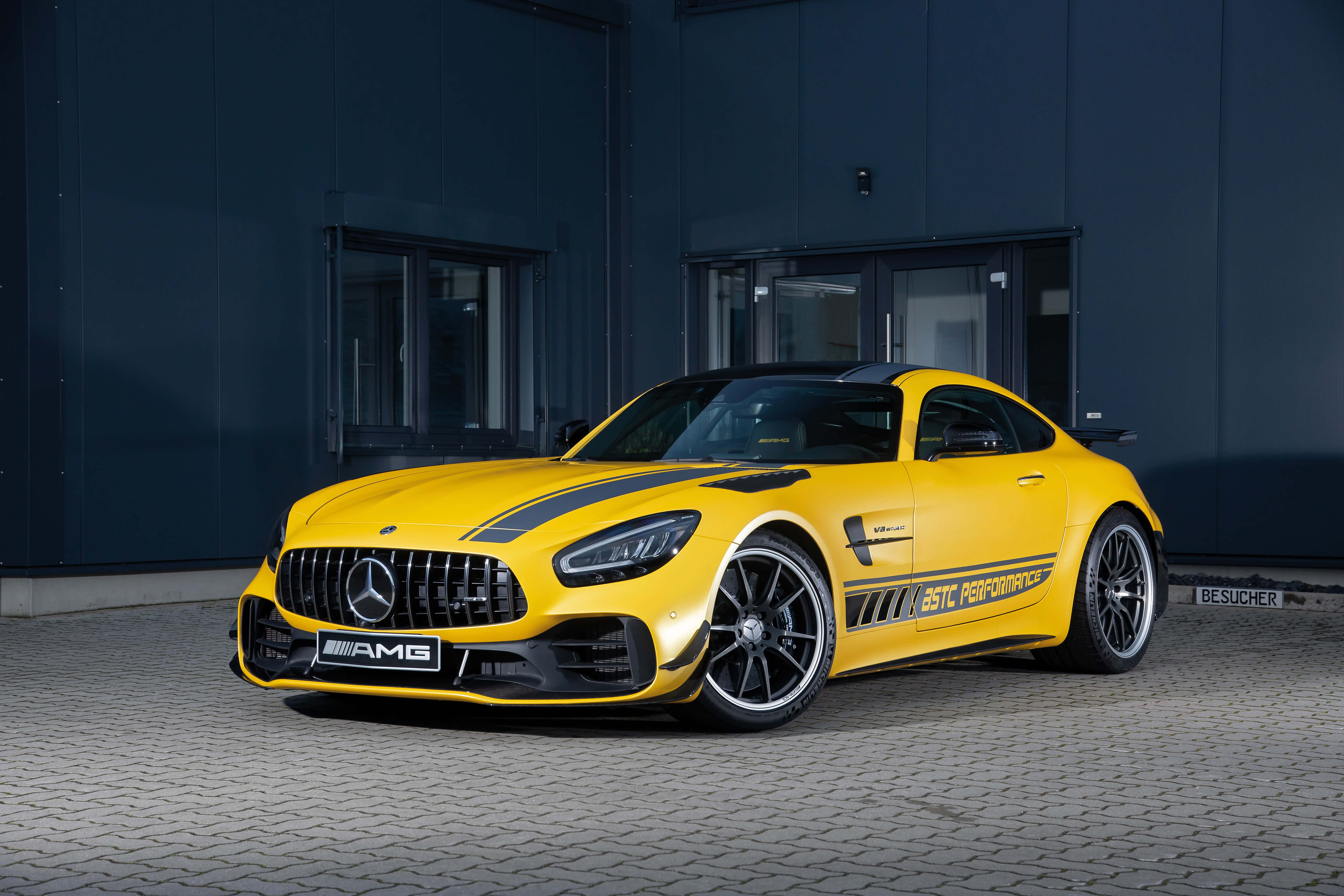 跑車 桌布 壁紙 — 一輛鮮黃色AMG GT R跑車帶有黑色賽車條紋，停放在鋪砌表面上，背景是深藍色工業建築。