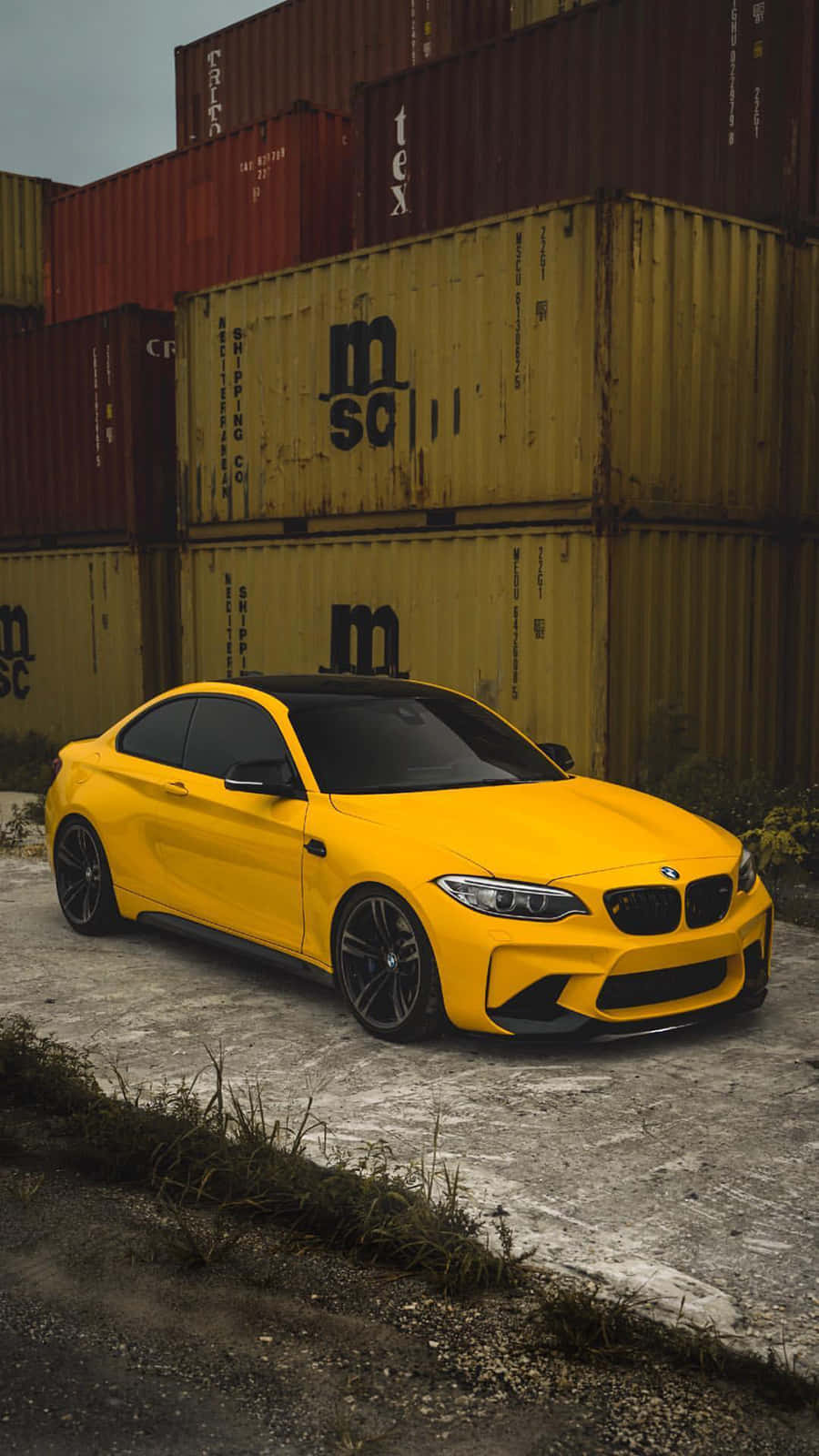 BMW 動圖 桌布 壁紙 — 一輛鮮黃色BMW M2雙門跑車停泊在裂開的混凝土上，旁邊是堆疊的貨運集裝箱，天空多雲。