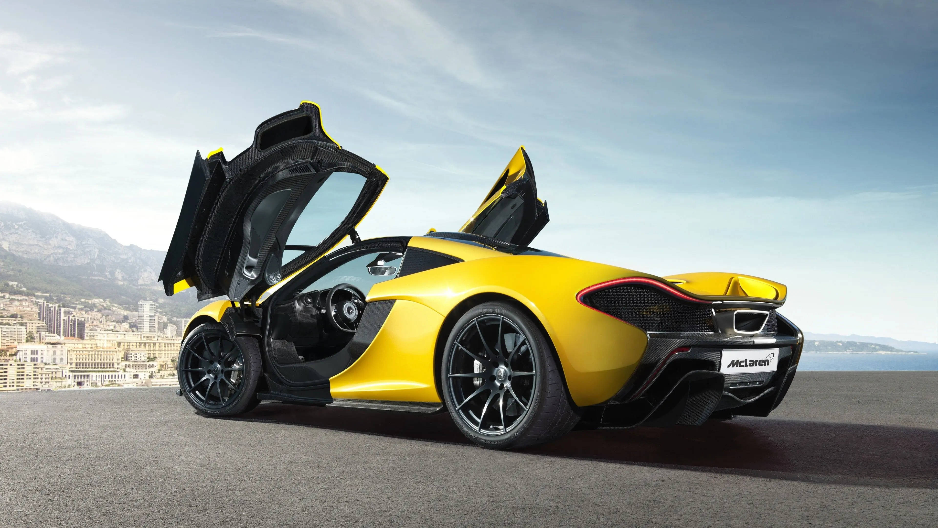 跑車 桌布 壁紙 — 一輛鮮黃色McLaren跑車，打開蝴蝶門，停泊在海岸懸崖上，俯瞰晴朗天空下的城市景觀。