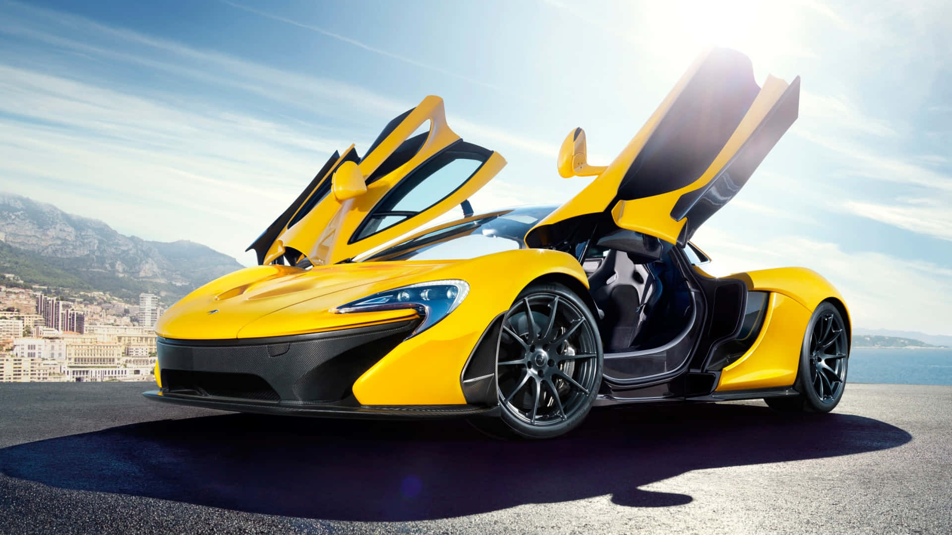 1920 X 1080 Car 桌布 壁紙 — 一輛鮮黃色McLaren跑車，鷗翼車門打開，停在沿海公路上，俯瞰城市與大海，天空湛藍明亮。