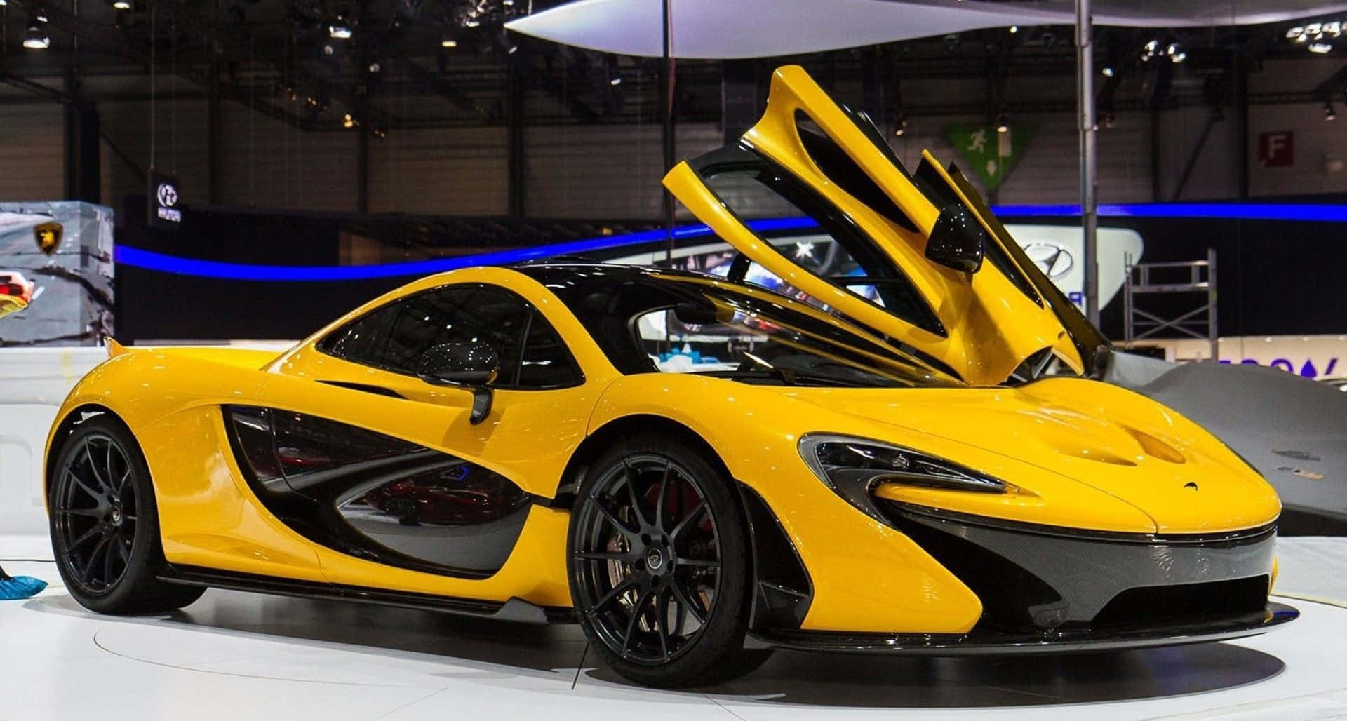 Top 10 Car 桌布 壁紙 — 一輛鮮黃色McLaren跑車，鷗翼門打開，在白色平台上明亮的展覽燈光下展示。