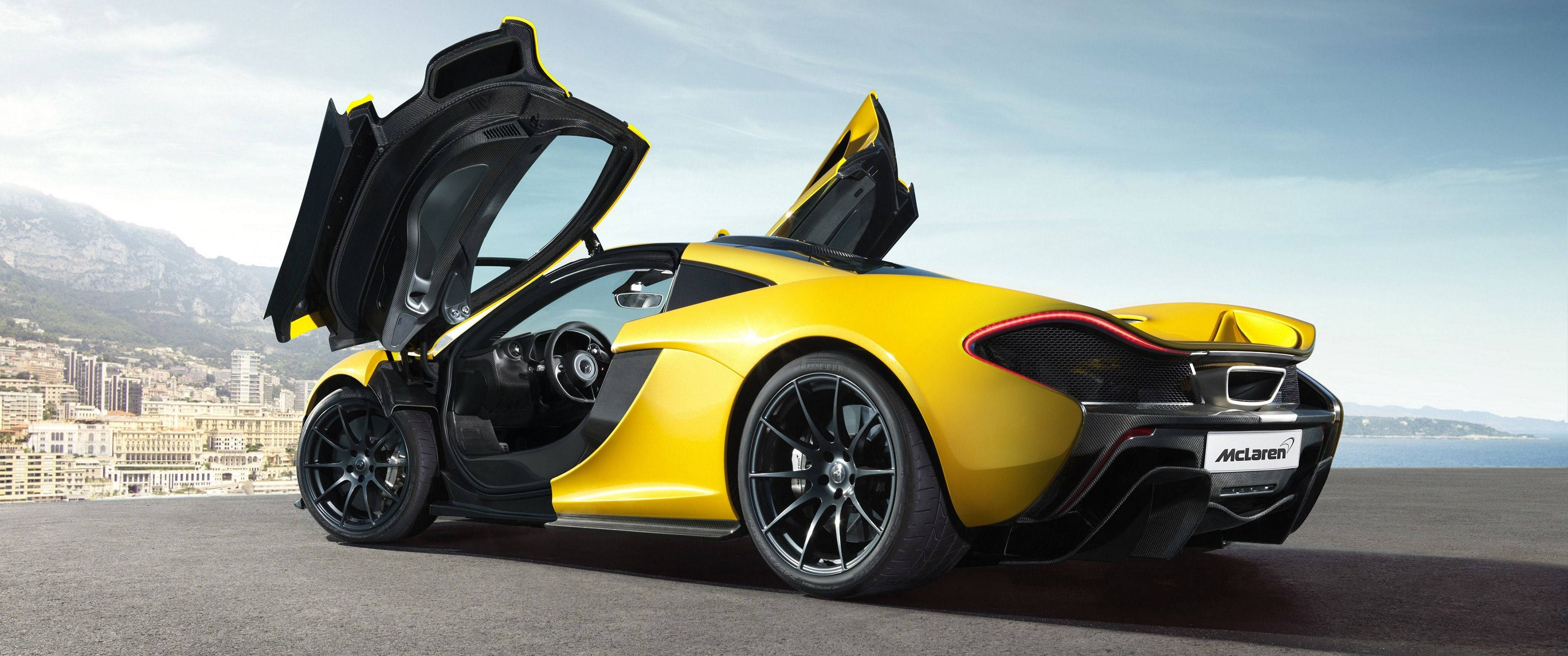 Cool Car 桌布 壁紙 — 一輛鮮黃色McLaren跑車，鷗翼車門打開，停在沿海公路上，俯瞰城市和山脈，天空晴朗。
