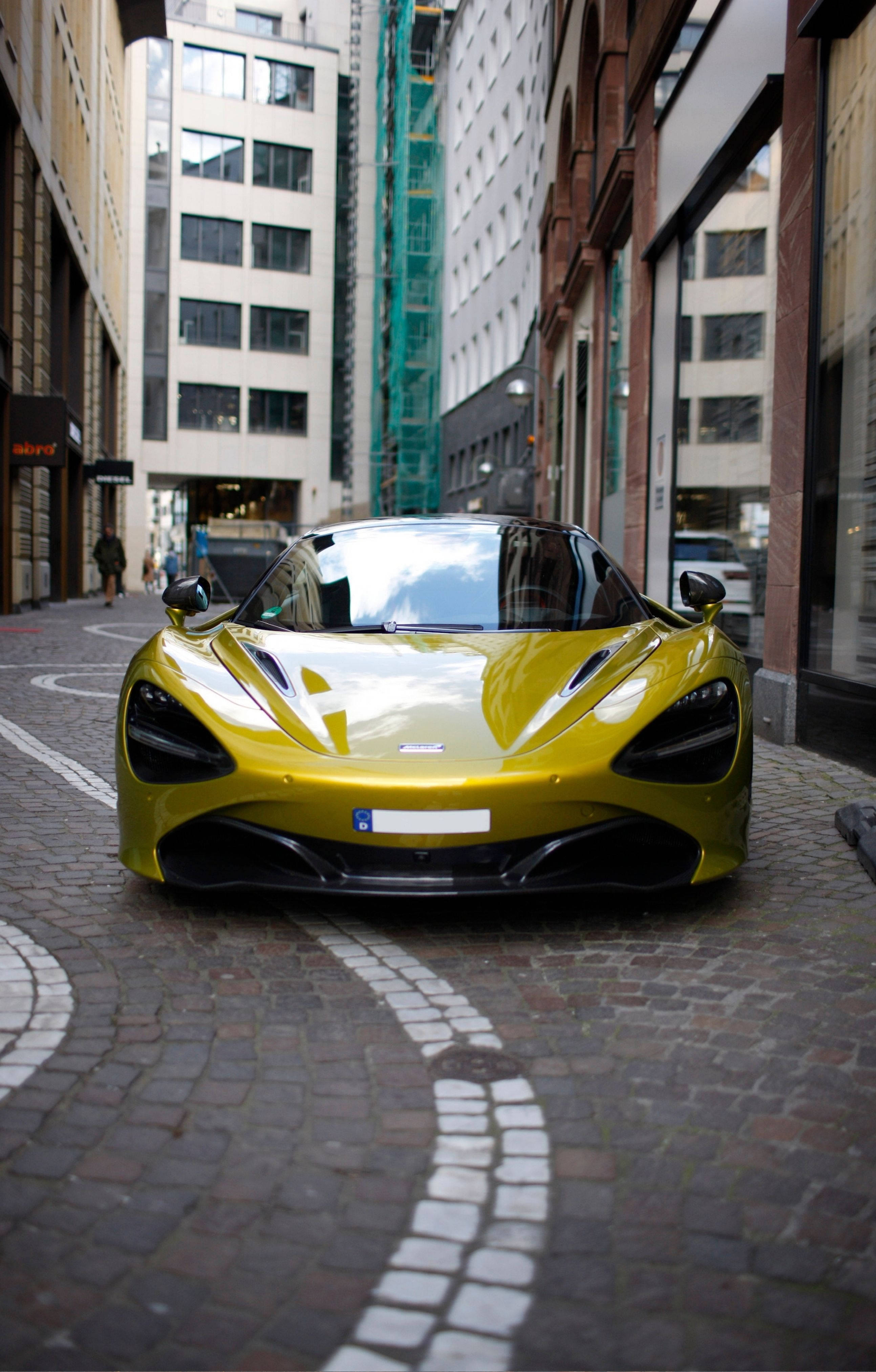 桌布 壁紙 — 一輛鮮黃色McLaren跑車停泊在鵝卵石街道上，兩旁是高聳的城市建築。