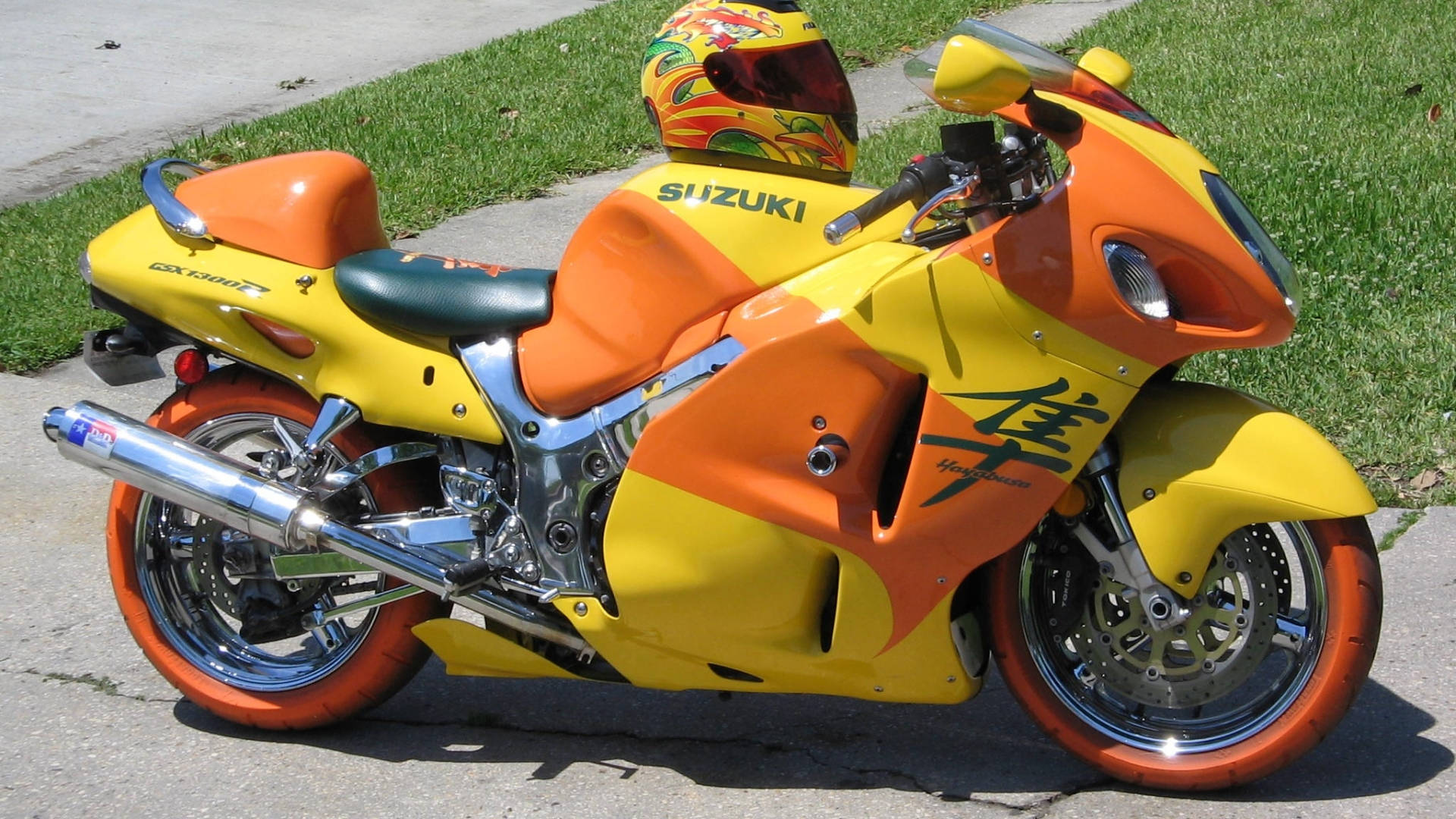 Suzuki 桌布 壁紙 — 一輛鮮黃色和橙色的鈴木隼機車停在人行道上，旁邊有綠草，在明亮的陽光下。