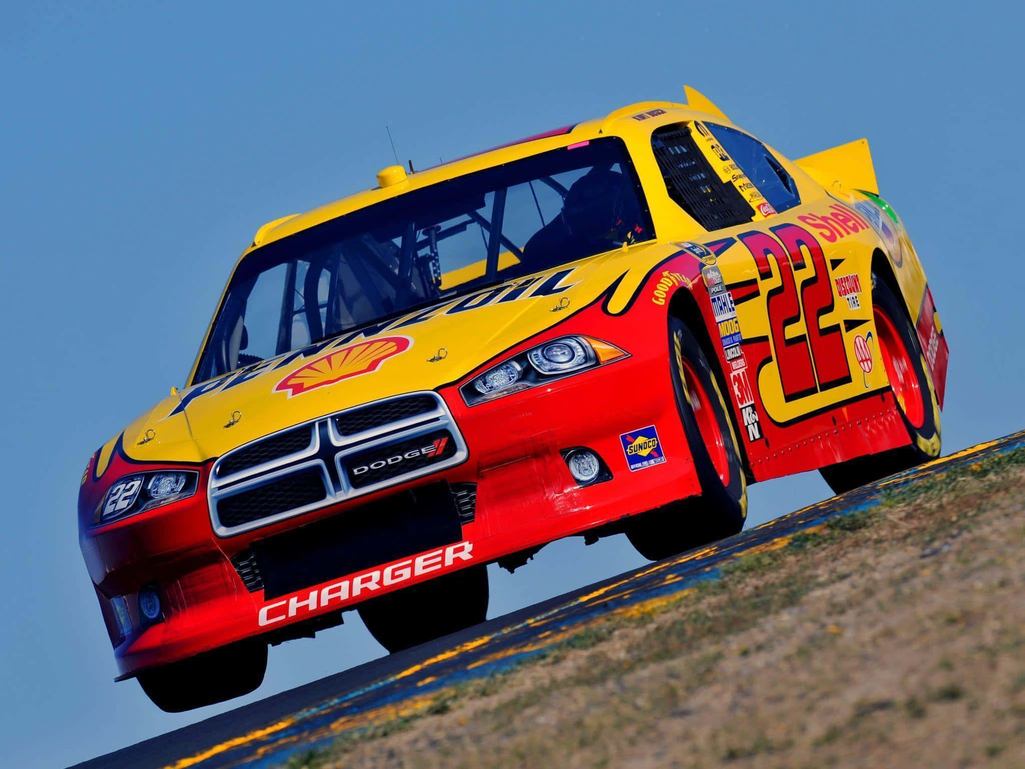 nascar 桌布 壁紙 — 一輛鮮黃色和紅色的道奇Charger賽車（編號22）在晴朗藍天下於賽道上空飛翔。