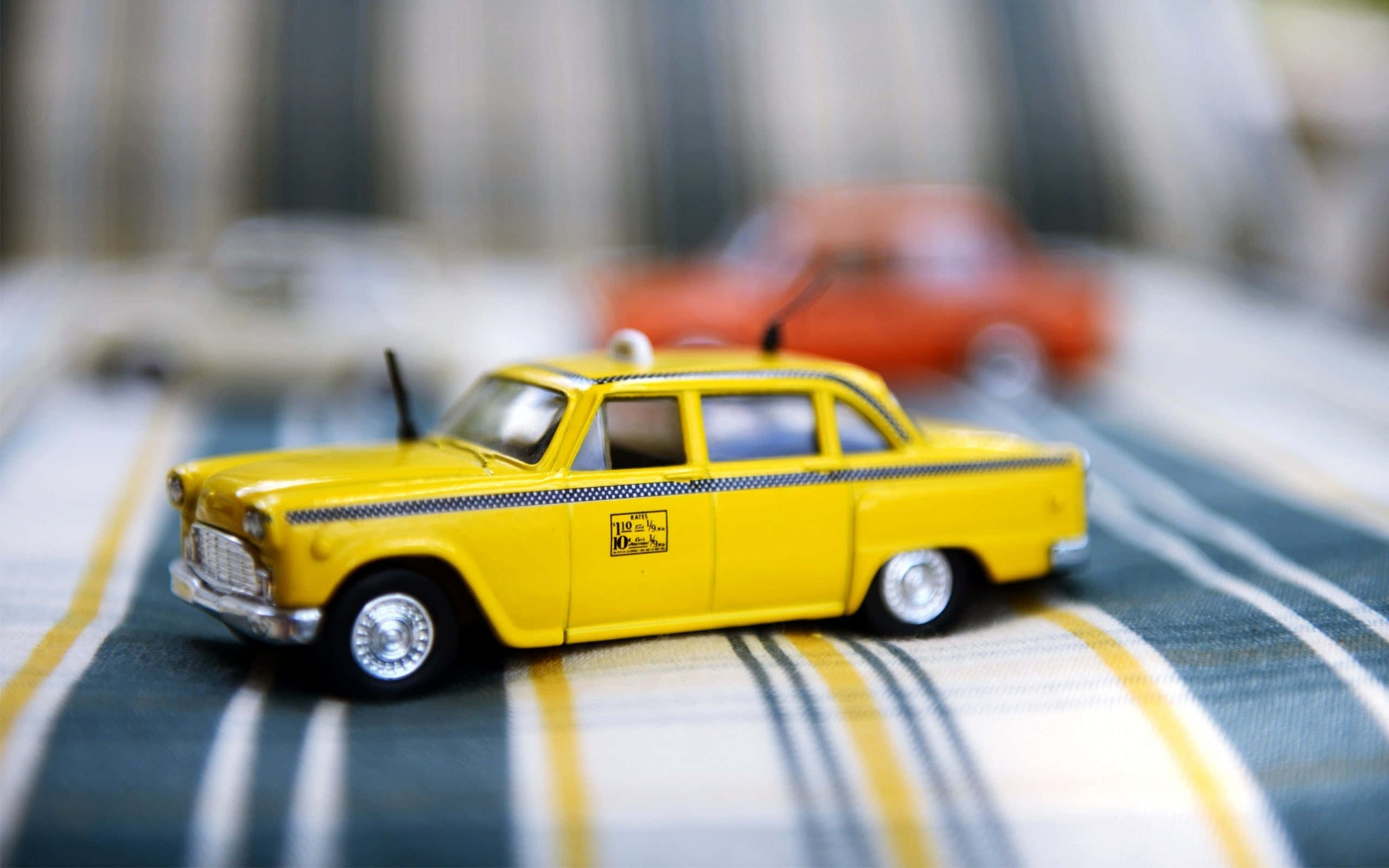 taxi 桌布 壁紙 — 一輛鮮黃色復古玩具計程車清晰對焦在條紋布料上，背景有模糊的玩具車。