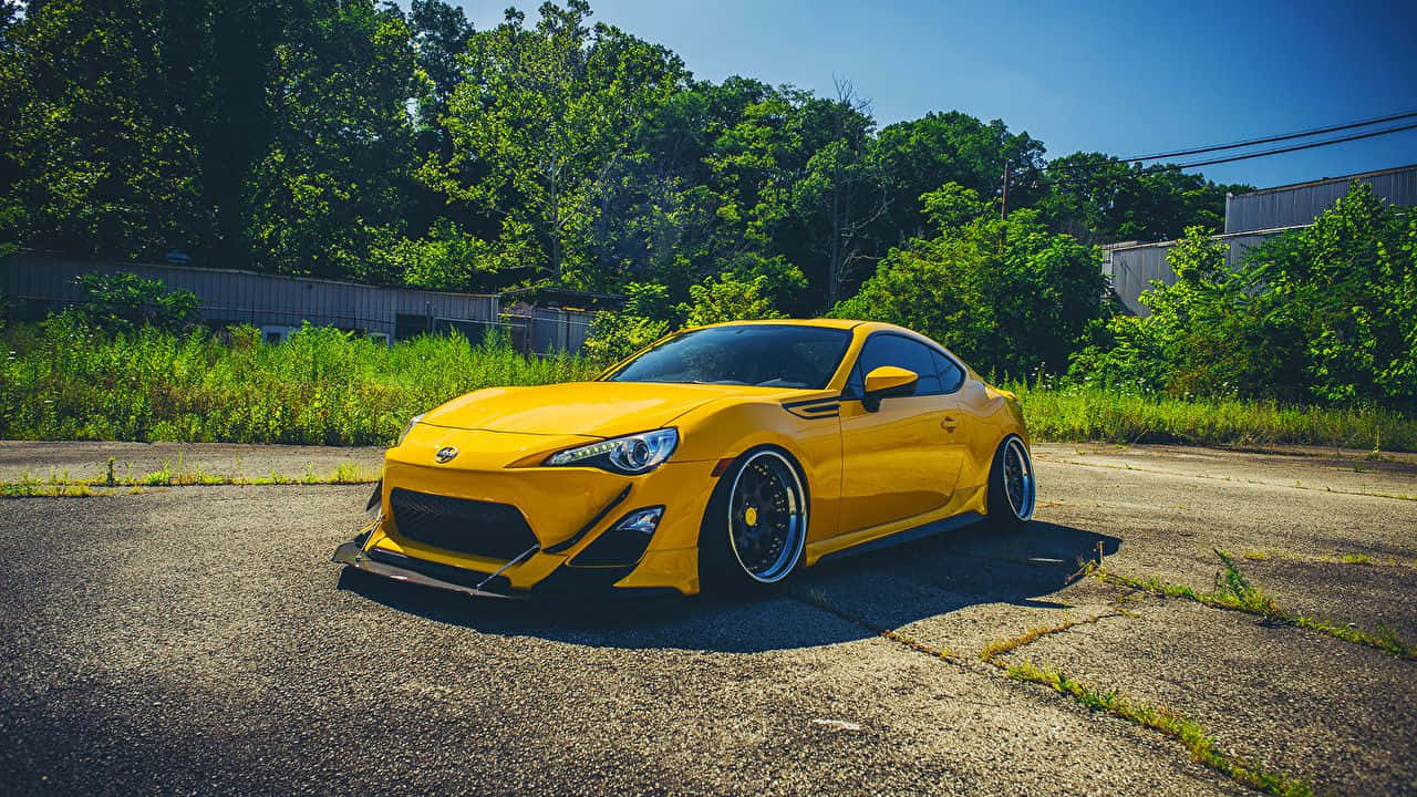 Yellow Car 桌布 壁紙 — 一輛鮮黃色改裝跑車停放在裂開的柏油路上，陽光燦爛，背景是茂密的綠樹。