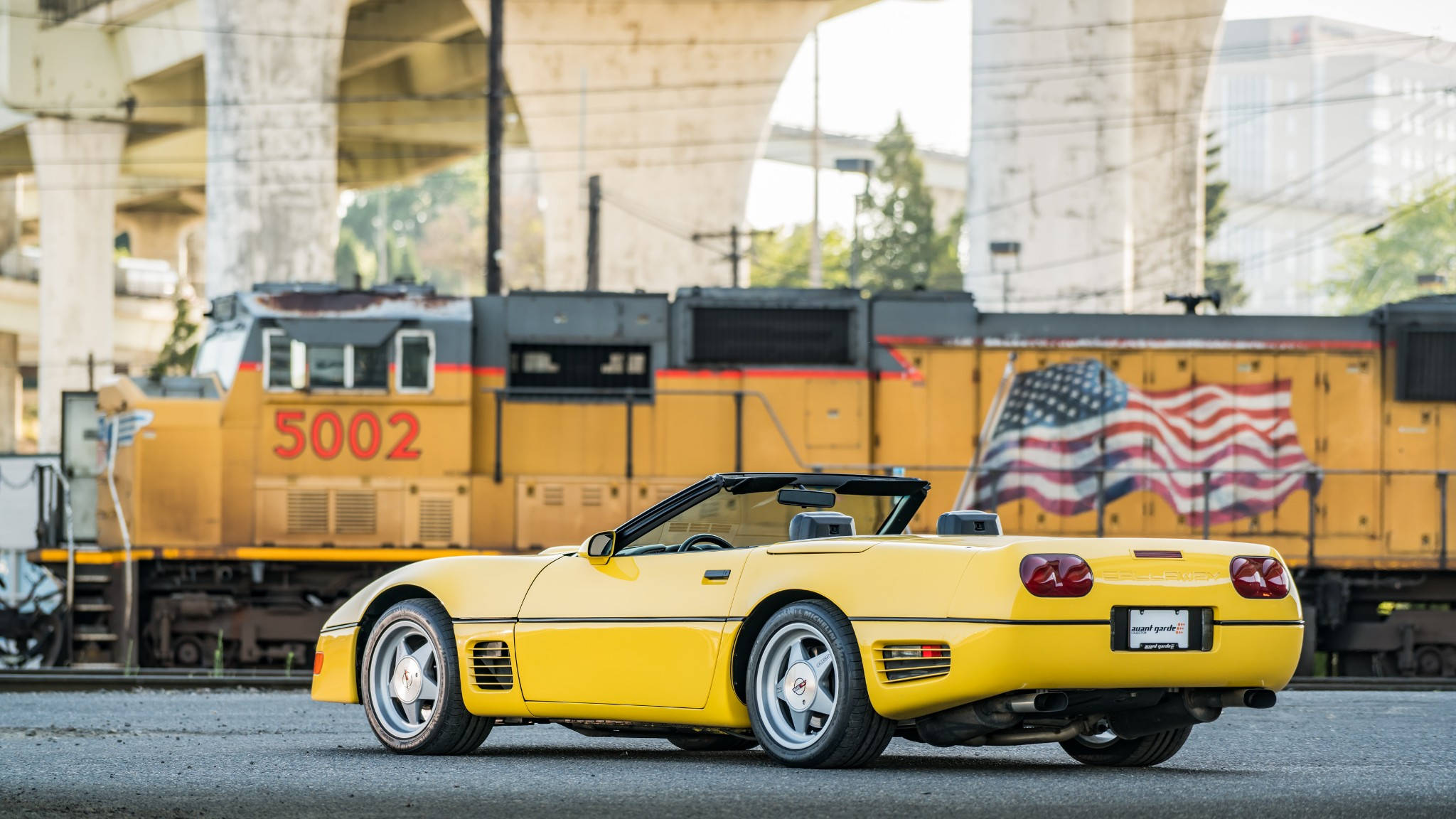 corvette 桌布 壁紙 — 一輛鮮黃色敞篷跑車停在高架橋下，背景有黃色火車和美國國旗。