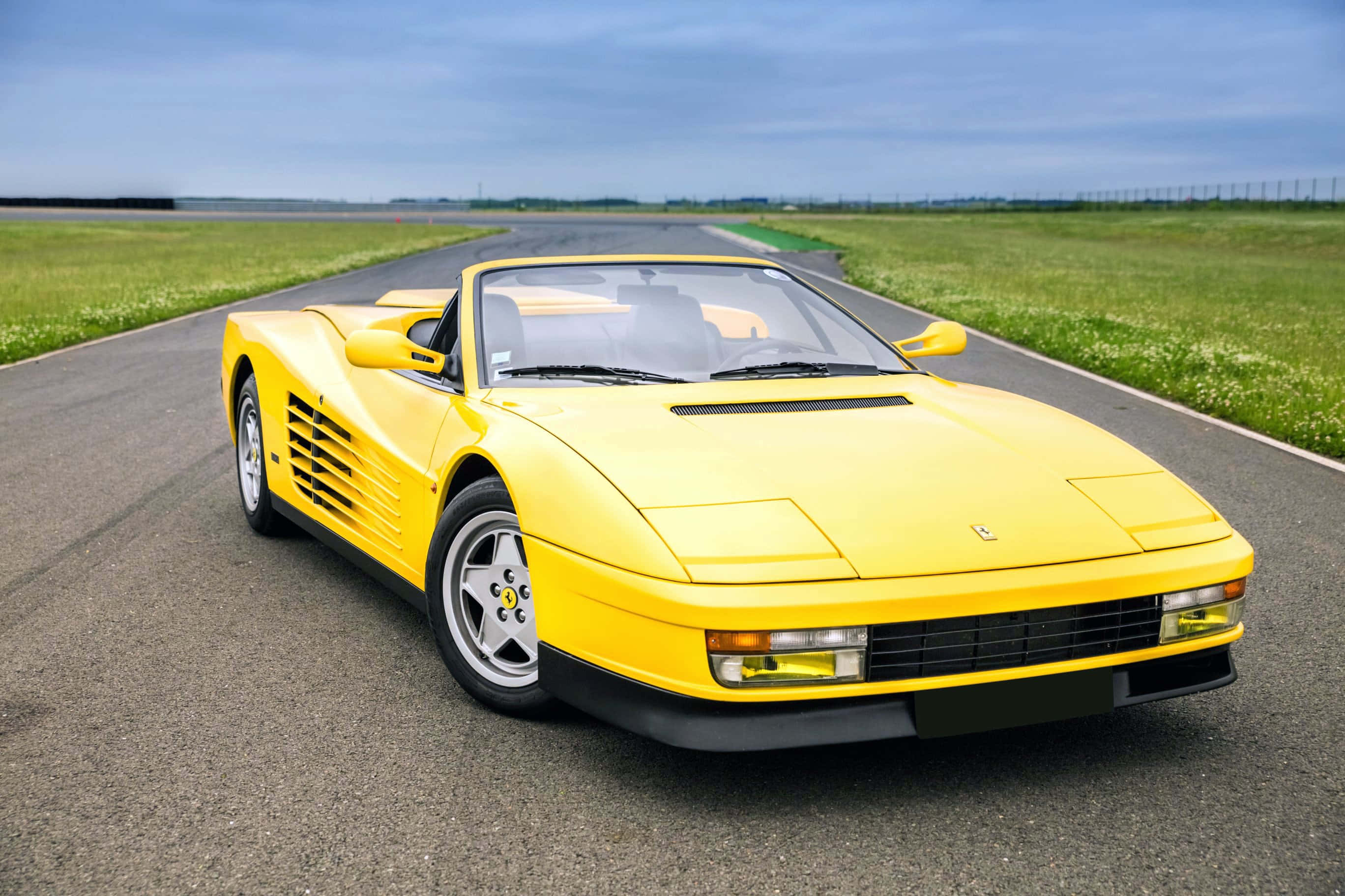 Ferrari Testarossa 桌布 壁紙 — 一輛鮮黃色法拉利Testarossa敞篷車停放在空曠的瀝青路上，兩旁是綠草，天空多雲。