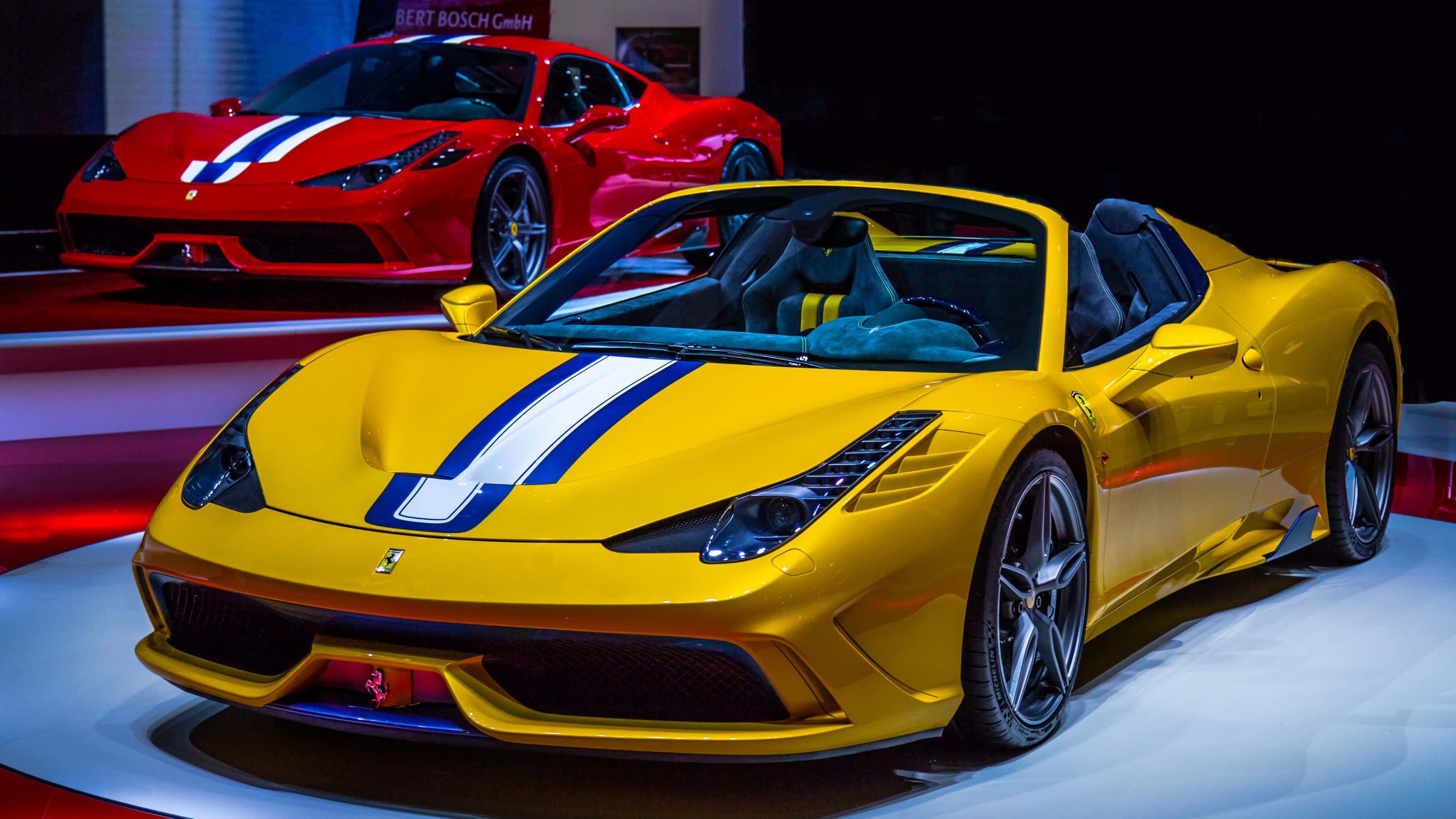 Ferrari 458 Speciale 桌布 壁紙 — 一輛鮮黃色法拉利敞篷車，帶有藍白賽車條紋，在明亮燈光下閃閃發光，停在一輛紅色法拉利前面，於車展中展出。