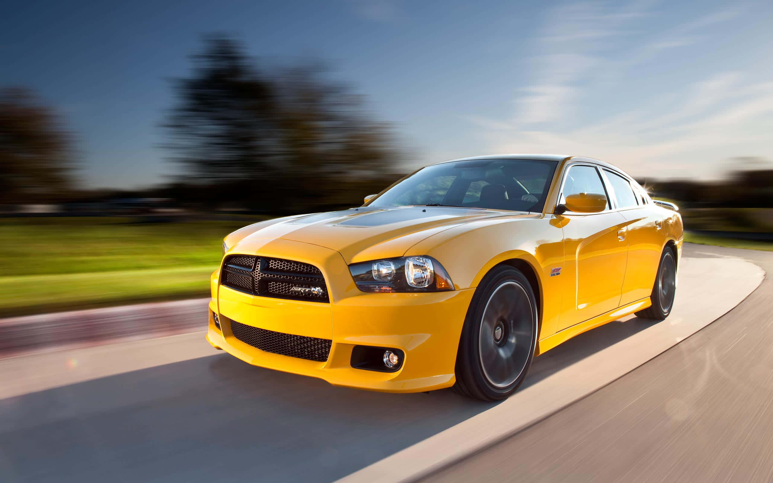 Yellow Car 桌布 壁紙 — 一輛鮮黃色的 Dodge Charger 沿著彎曲的道路疾馳，背景是模糊的綠樹和天空。