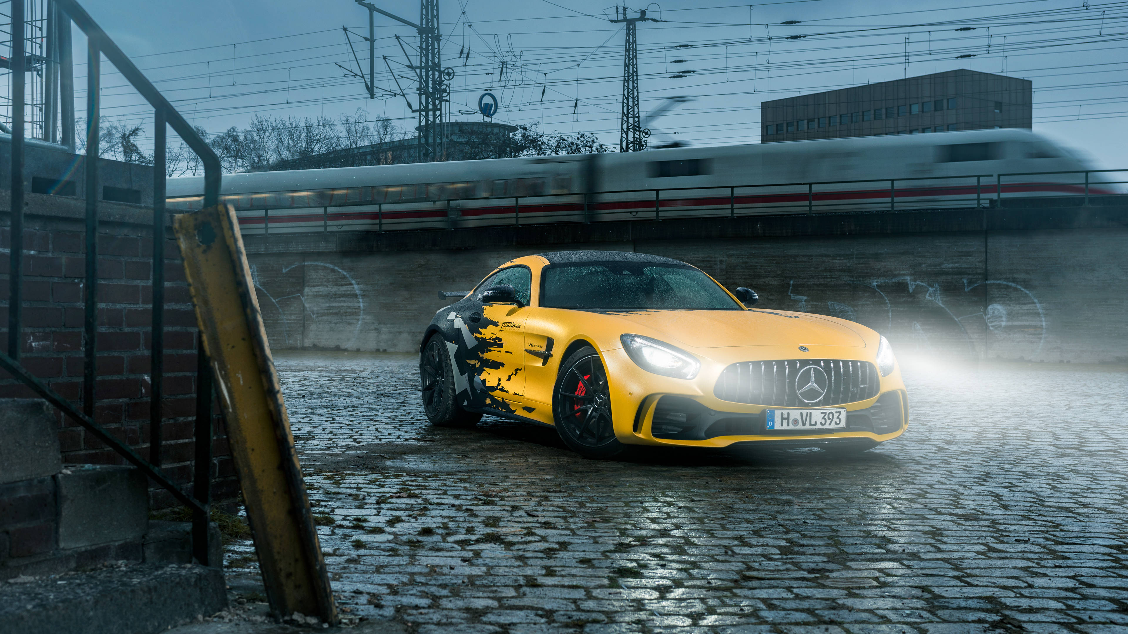 Amg 桌布 壁紙 — 一輛鮮黃色的 Mercedes-AMG GT 跑車停在濕漉漉的鵝卵石上，一列高速列車模糊地從上方駛過。