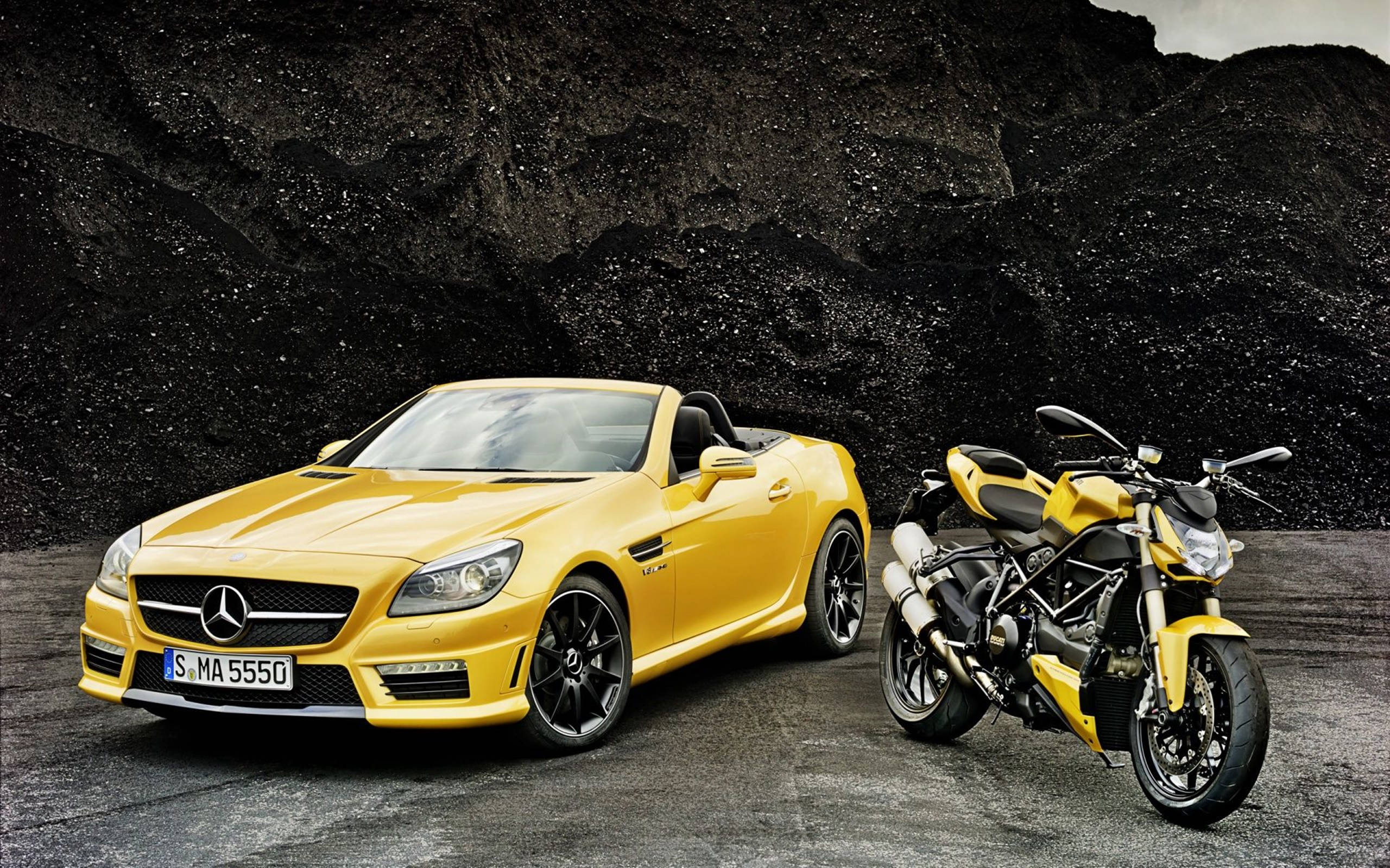 高清 桌布 壁紙 — 一輛鮮黃色的 Mercedes SLK 敞篷車和一輛相配的黃色 Ducati 摩托車並排停放在深色紋理岩石牆前。