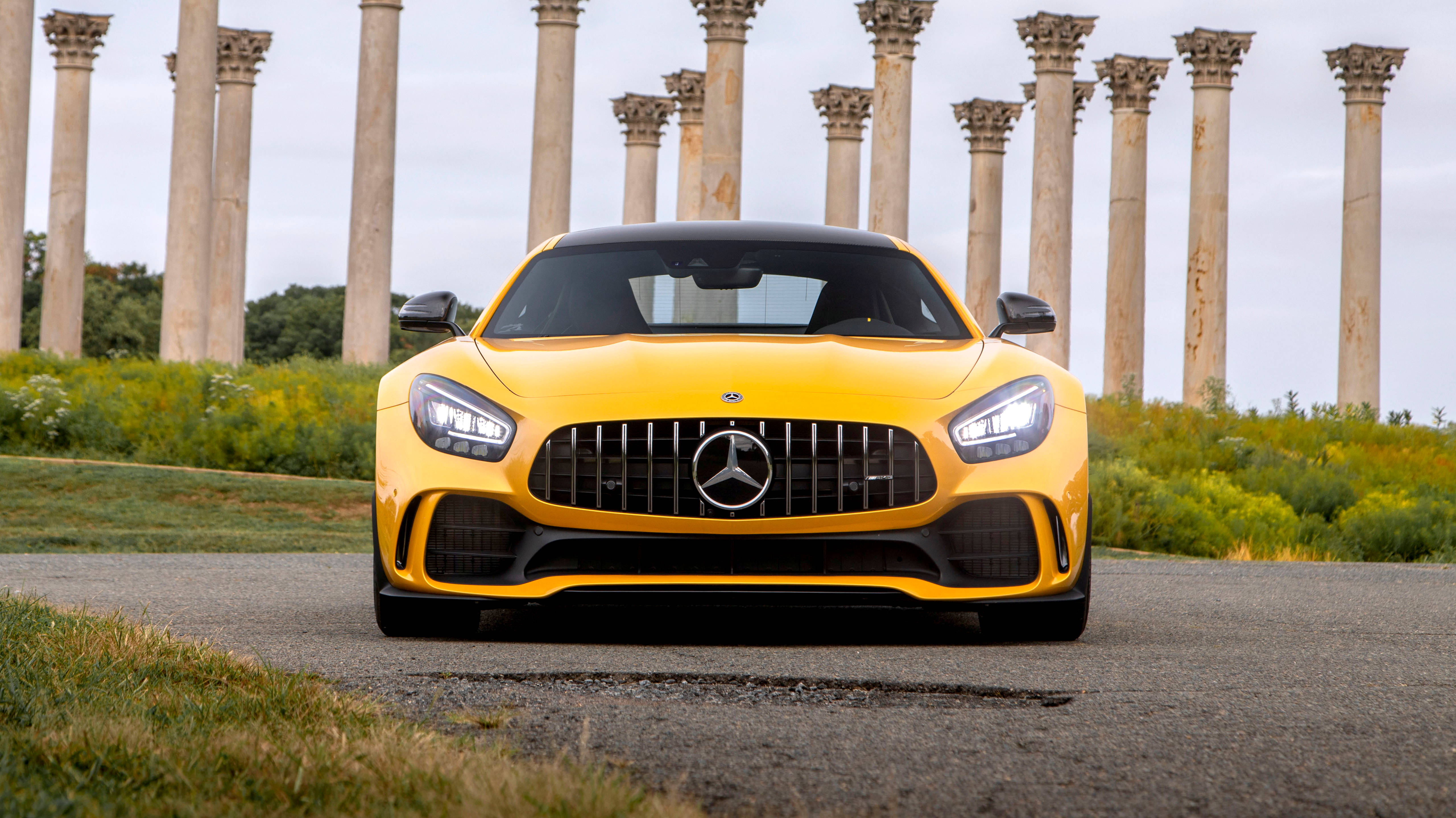 4K 奔馳壁紙 桌布 壁紙 — 一輛鮮黃色的 Mercedes-AMG GT 跑車在陰天之下，面向前方，停泊在由古老石柱環繞的鋪砌小徑上。