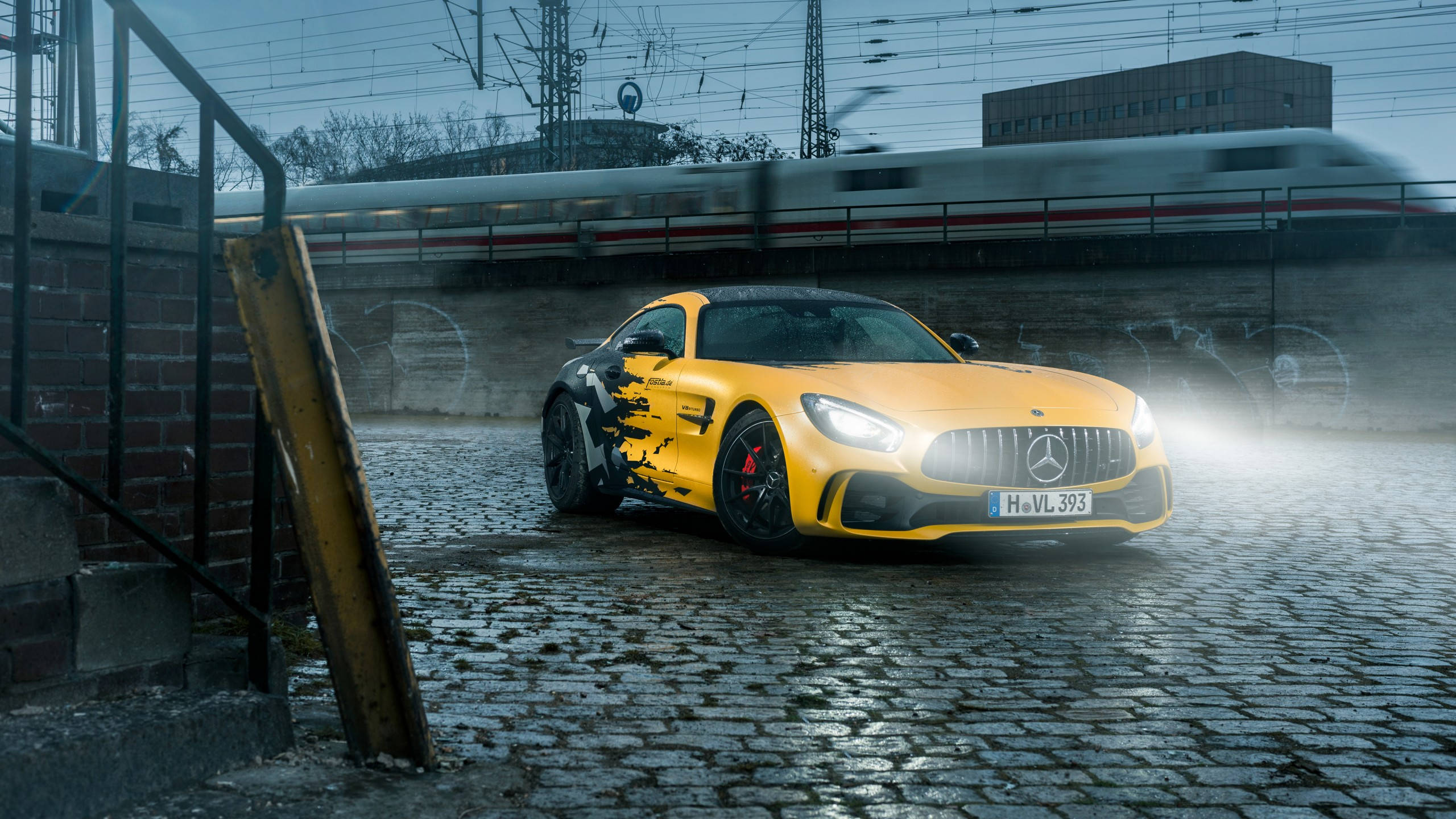Gtr 桌布 壁紙 — 一輛鮮黃色的 Mercedes-AMG GT 跑車在潮濕的鵝卵石平台上，於車頭燈下閃閃發光，一列模糊的高速列車從上方駛過。