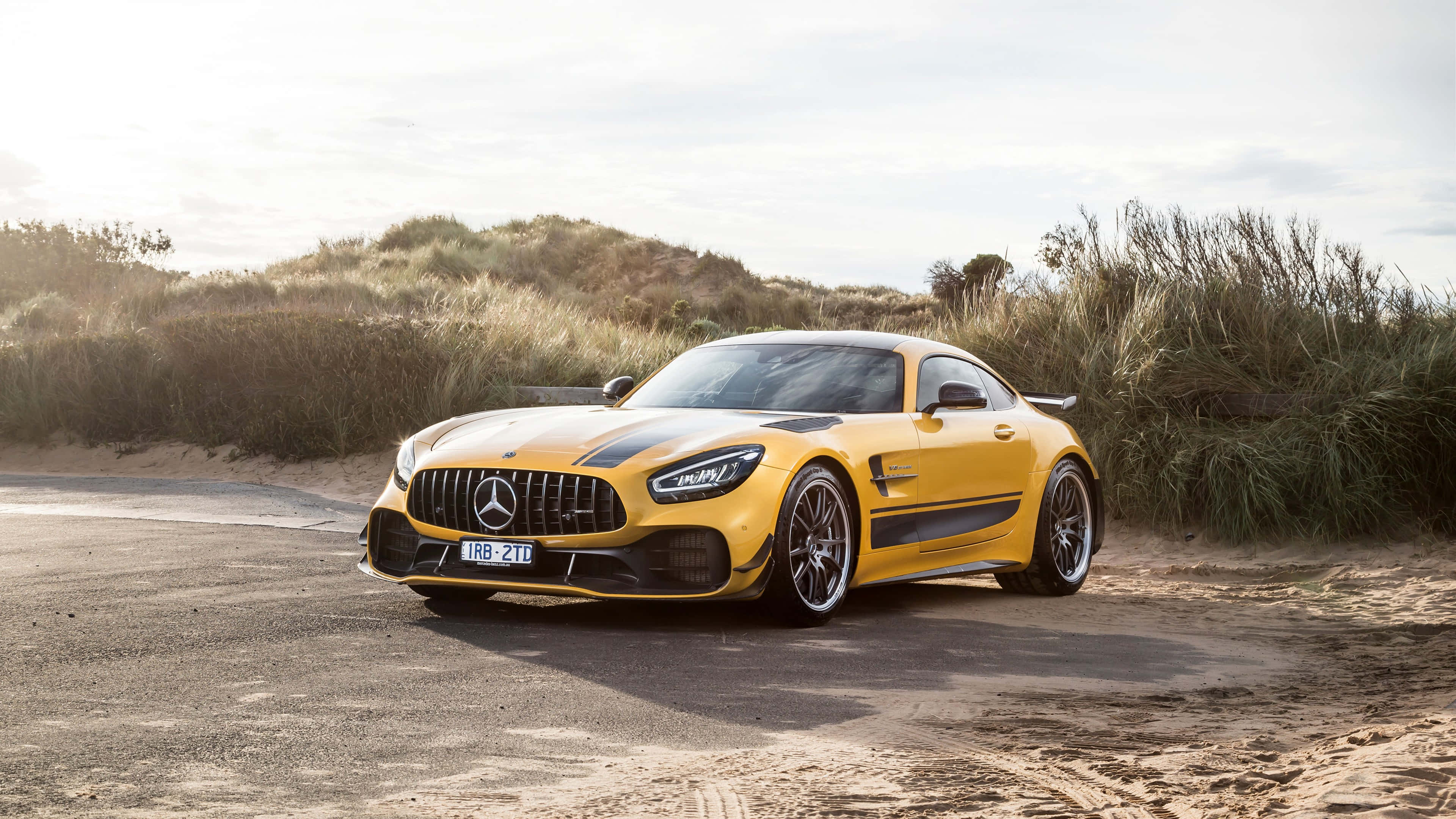 Mercedes 桌布 壁紙 — 一輛鮮黃色的 Mercedes-AMG GT Black Series 跑車停在沙路上，旁邊有沙丘草，在柔和日光下。