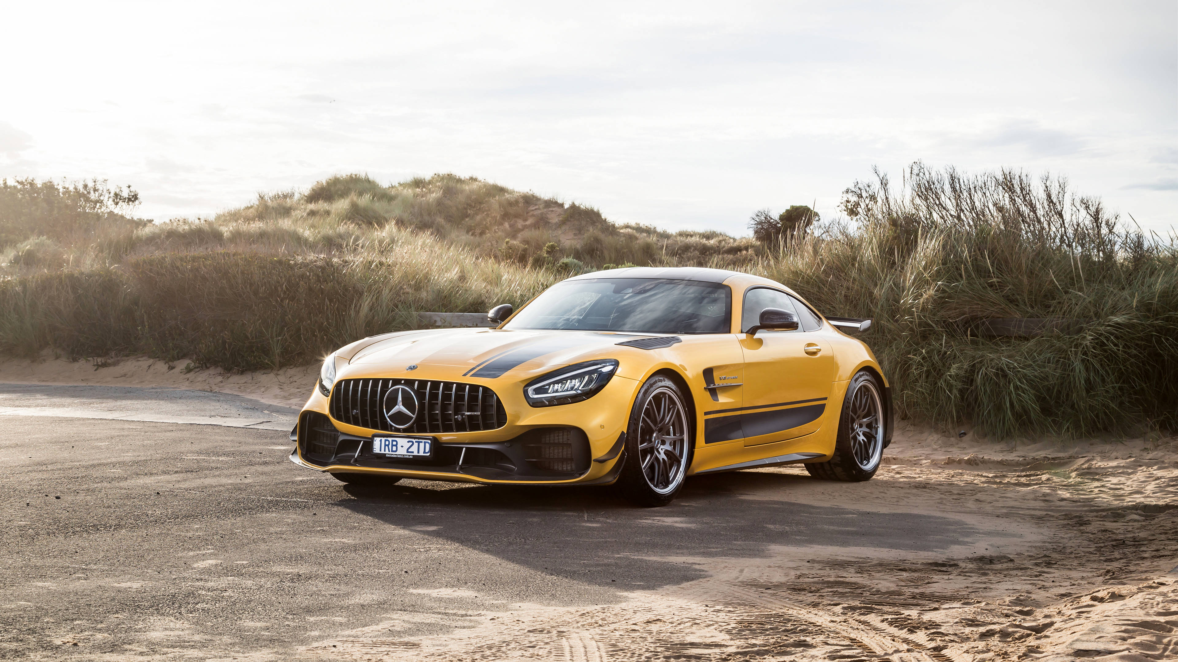 Amg 桌布 壁紙 — 一輛鮮黃色的 Mercedes-AMG GT Black Series 跑車停泊在沙路旁，旁邊是長滿草的沙丘，天空晴朗。