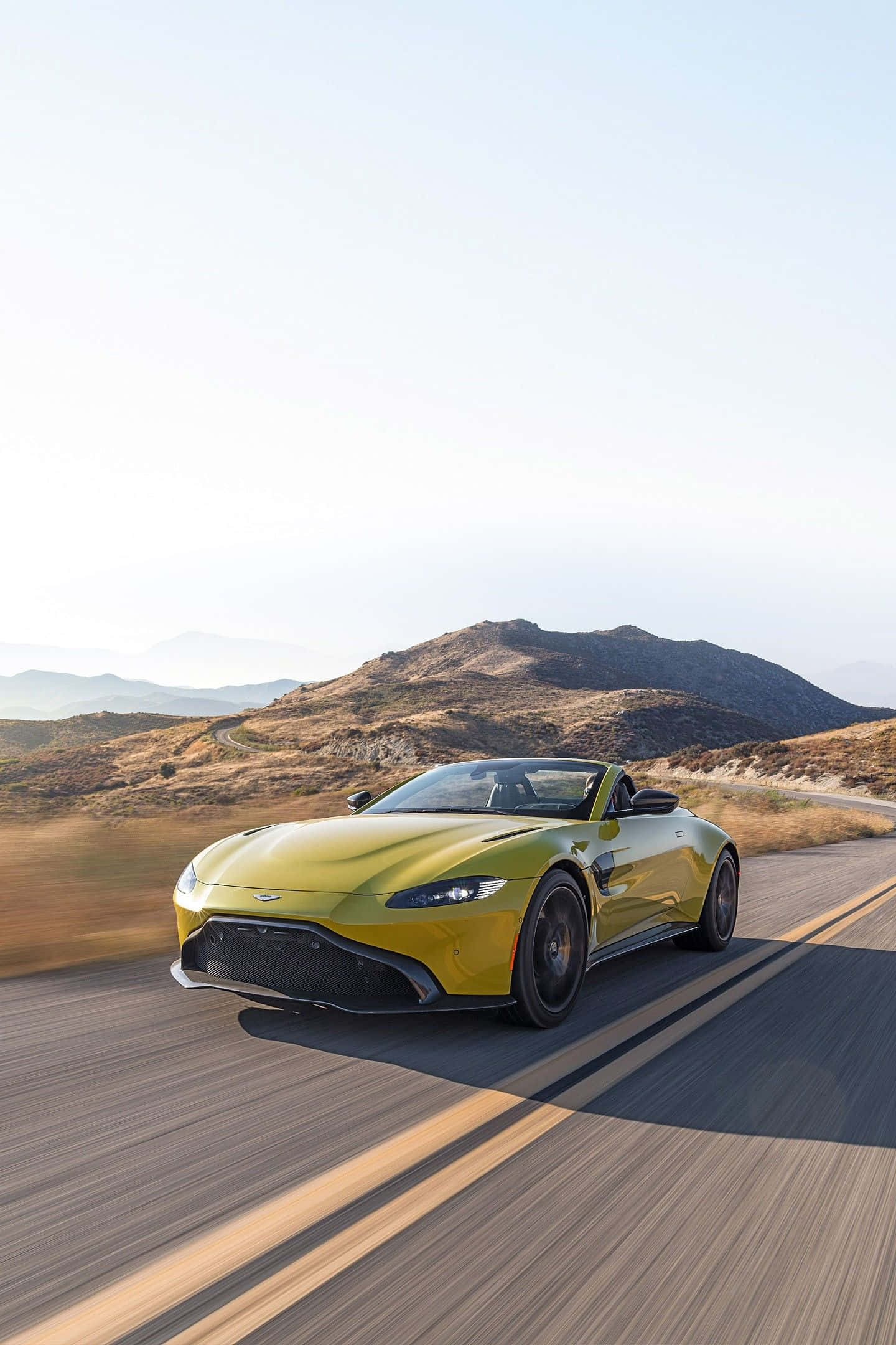 奧斯頓馬丁 桌布 壁紙 — 一輛鮮黃色的Aston Martin Vantage敞篷跑車在晴朗的天空下沿著蜿蜒的山路奔馳。