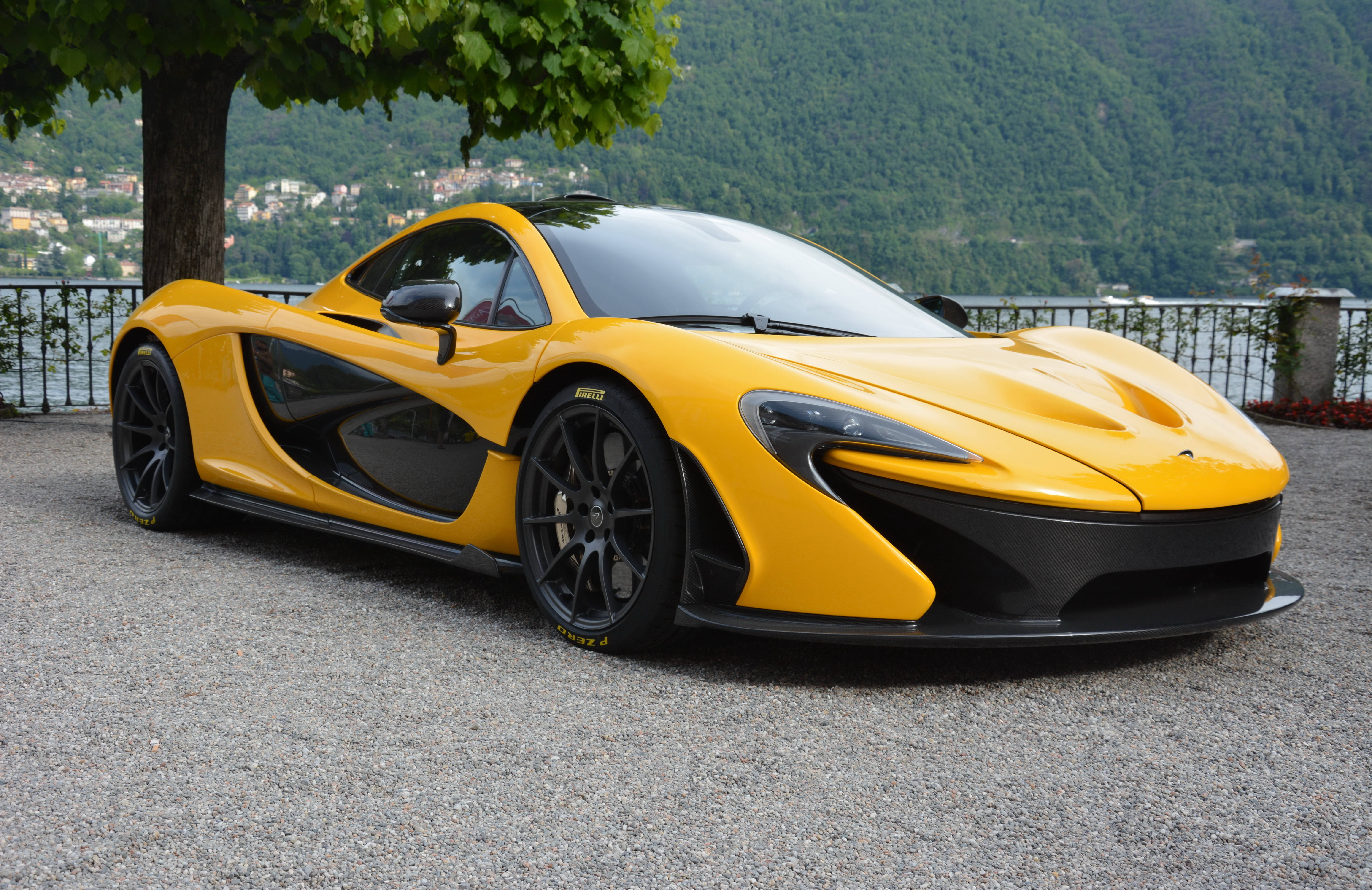 麥拉倫 桌布 壁紙 — 一輛鮮黃色的McLaren P1超級跑車停在碎石路上，旁邊有樹，背景是風景如畫的湖泊和山脈。