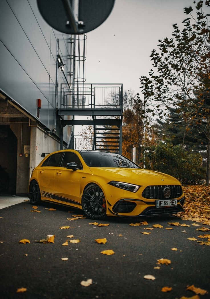 Hatchback 桌布 壁紙 — 一輛鮮黃色的Mercedes-AMG A45停放在一個佈滿落葉的瀝青停車場，旁邊是一座帶有金屬樓梯的現代建築。