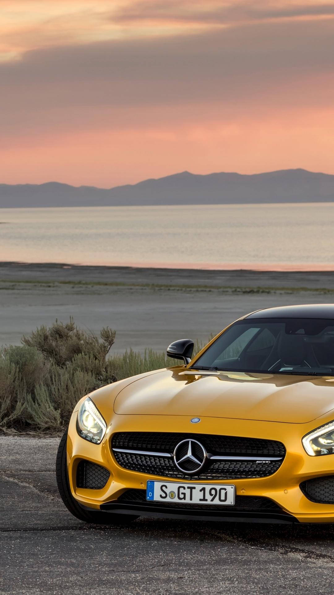 amg iPhone 桌布 壁紙 — 一輛鮮黃色的Mercedes-AMG GT停泊在日落時分的沿海公路上，背景有山脈和寧靜的大海。