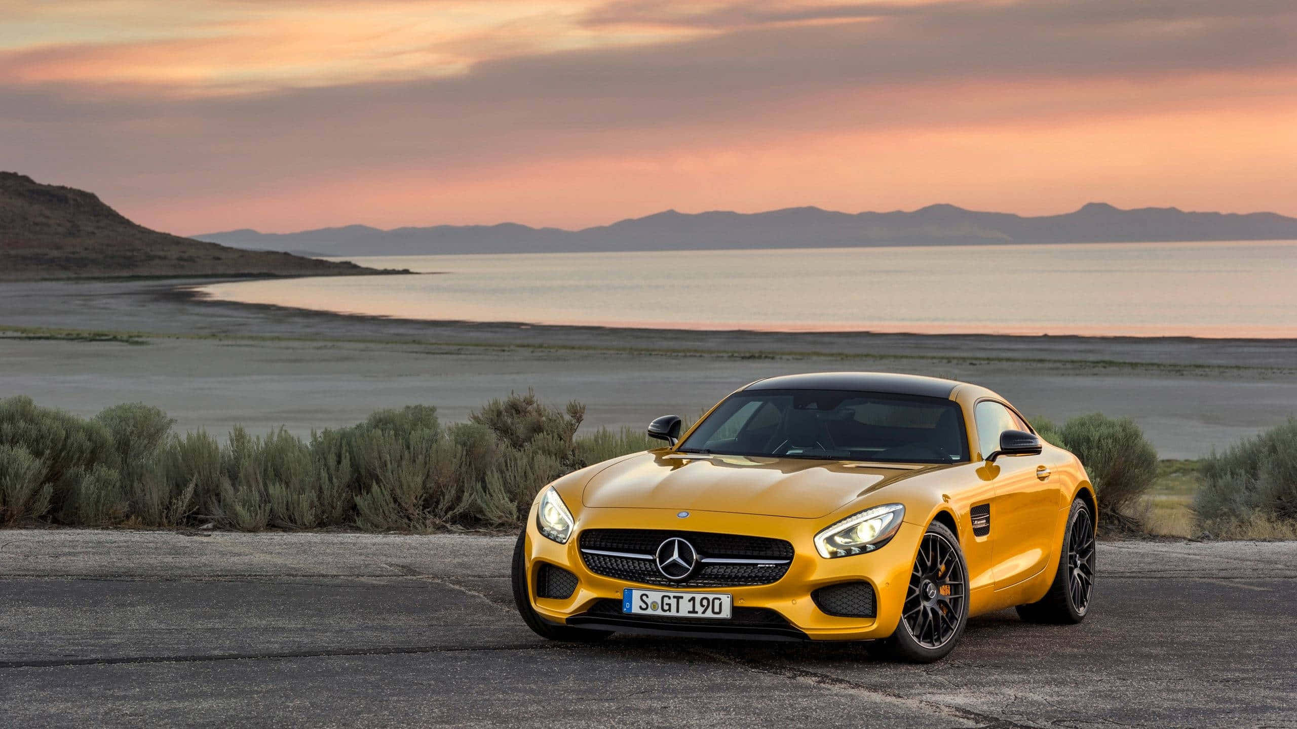 Gold Cars 桌布 壁紙 — 一輛鮮黃色的Mercedes-AMG GT停泊在日落時分的海岸公路上，背景有山脈和寧靜的水面。
