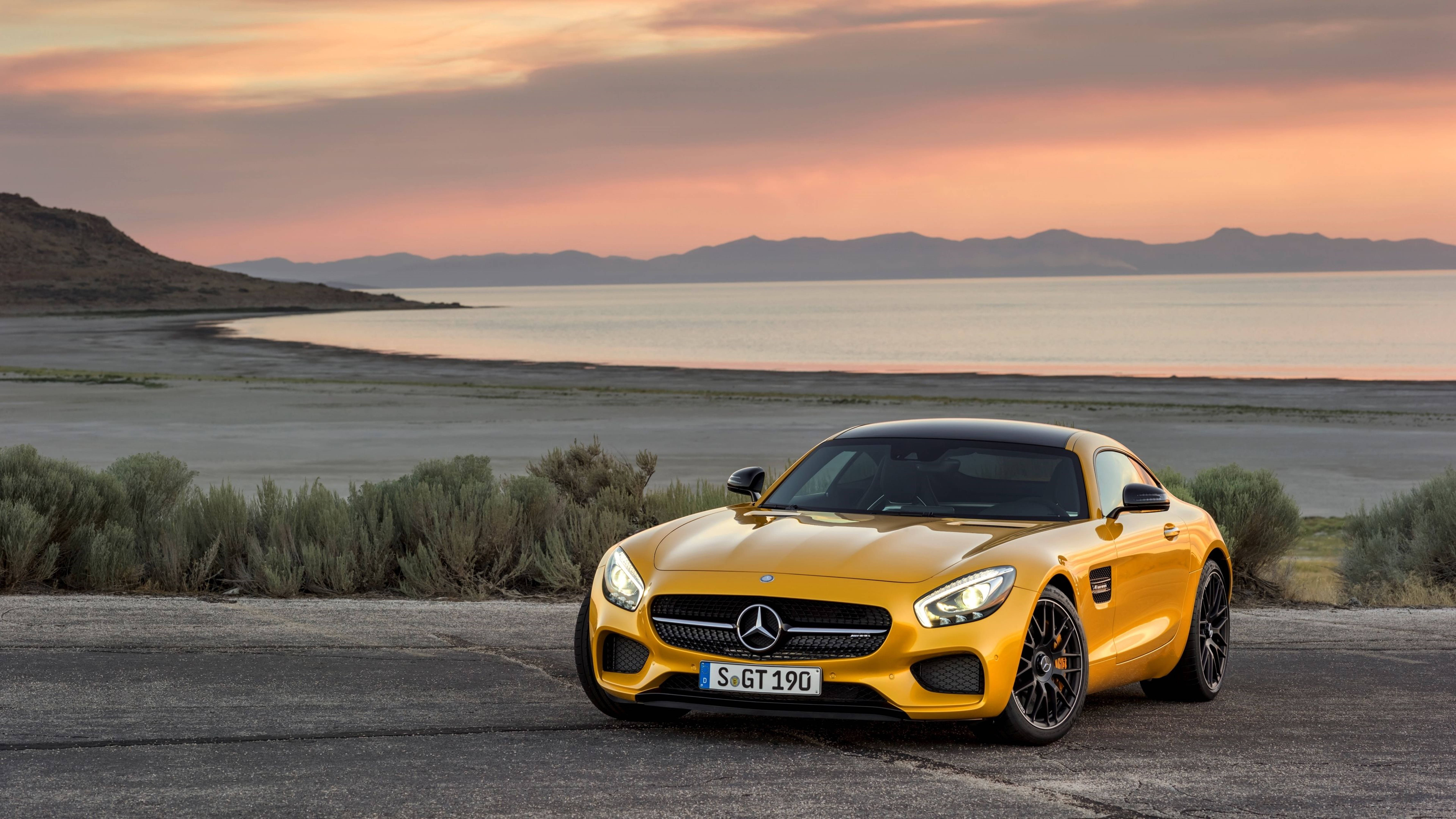 Mercedes 桌布 壁紙 — 一輛鮮黃色的Mercedes-AMG GT停泊在日落時分的海岸公路上，背景有山脈和寧靜的水面。