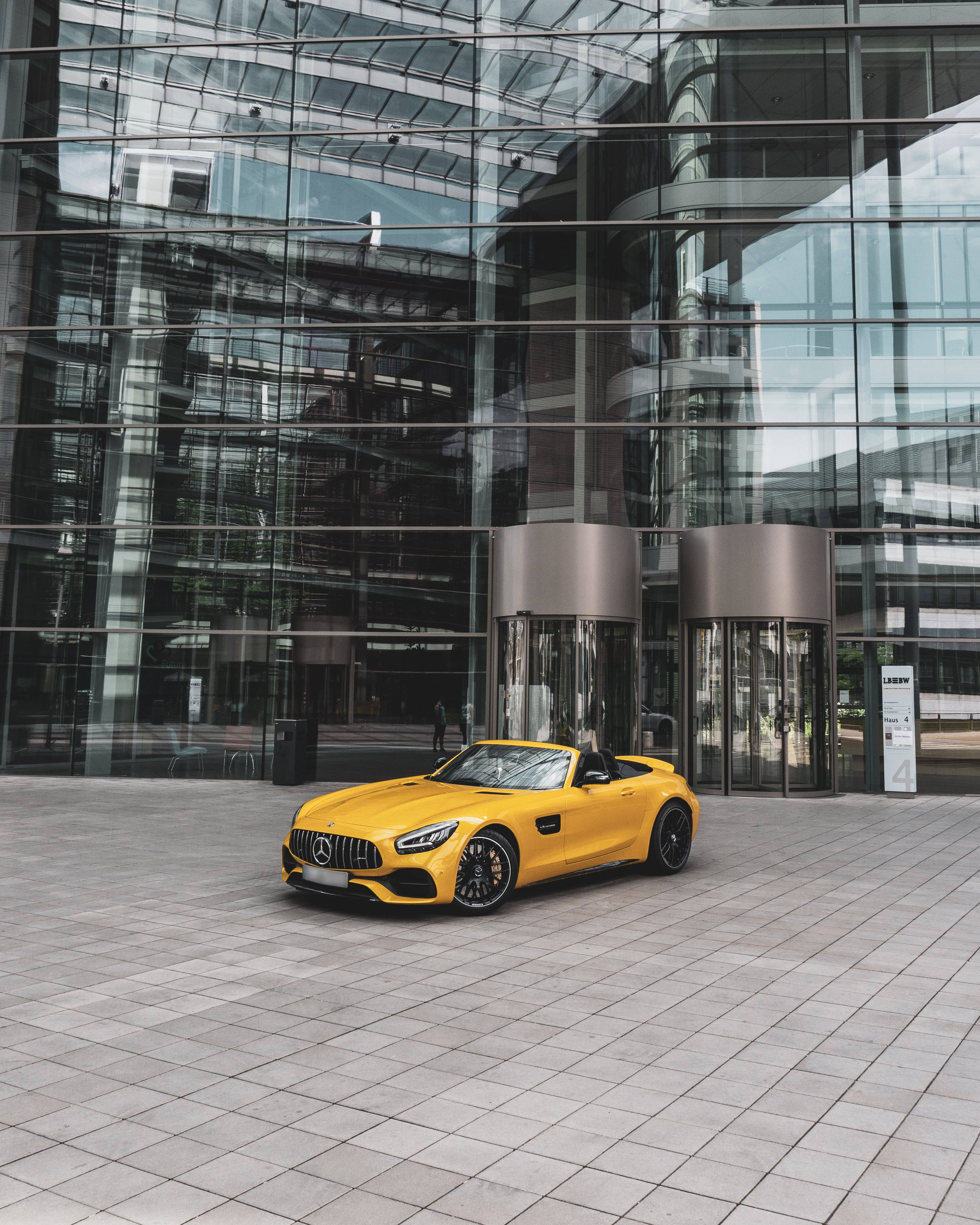 mercedes amg iPhone 桌布 壁紙 — 一輛鮮黃色的Mercedes-AMG GT Roadster停放在現代玻璃建築前的瓷磚廣場上。
