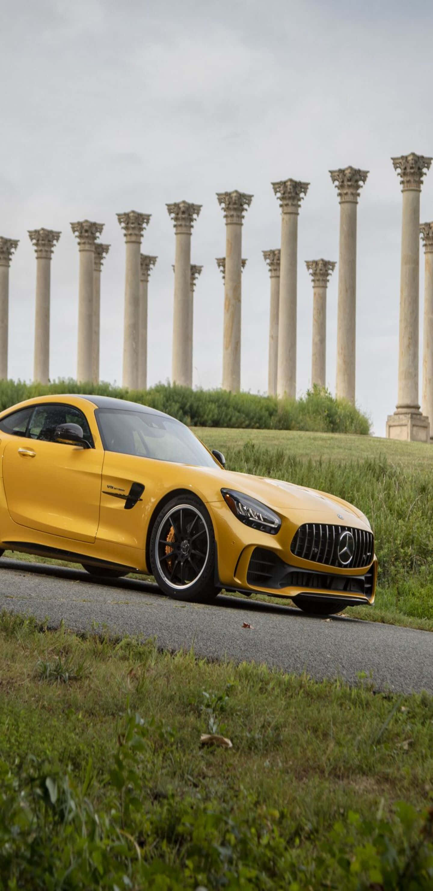Cool Car 桌布 壁紙 — 一輛鮮黃色的Mercedes-AMG GT跑車停在一條路上，前方是古老的石柱，天空多雲。