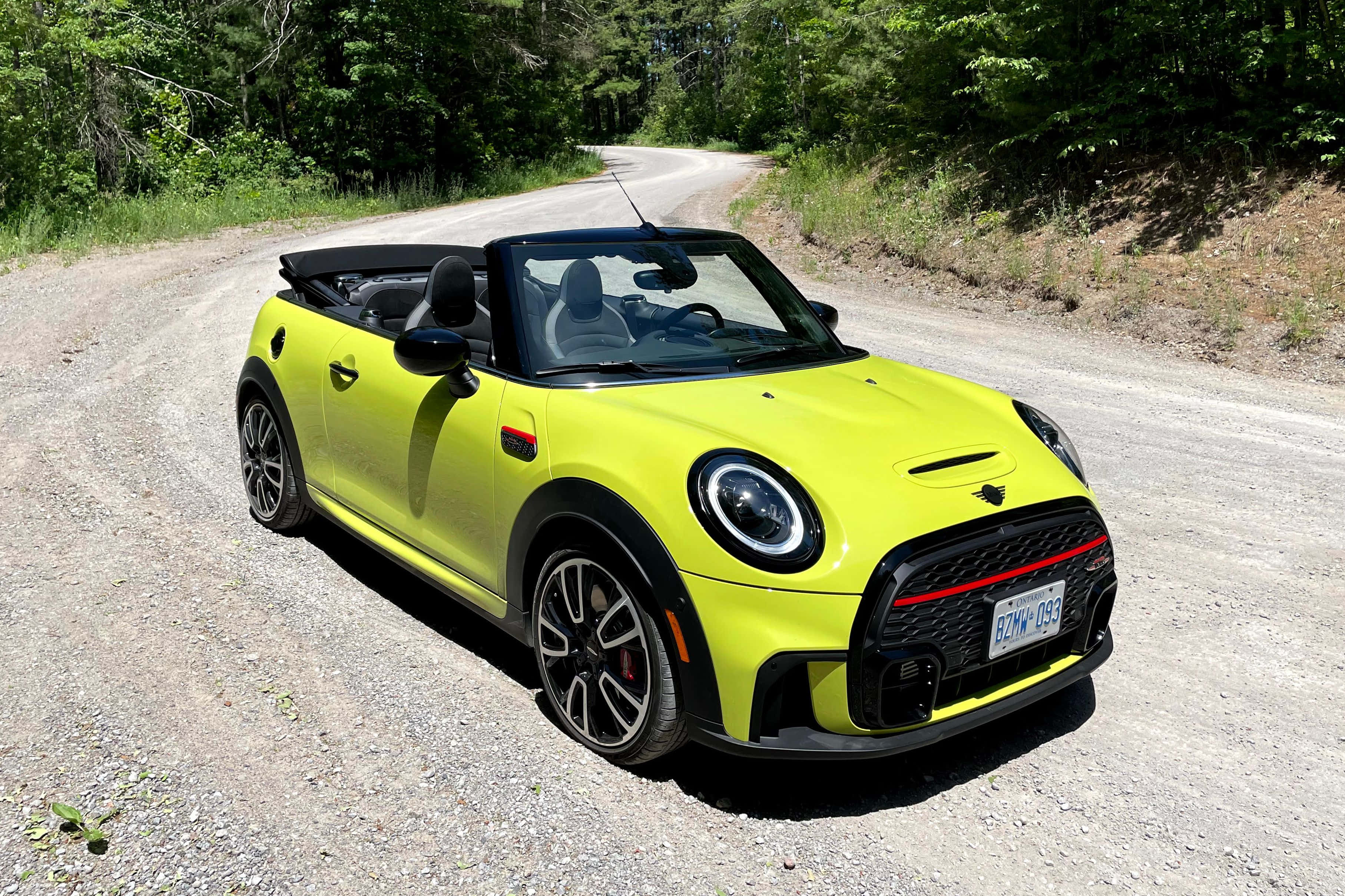 Mini Cooper Convertible 桌布 壁紙 — 一輛鮮黃色的MINI Cooper敞篷車停放在陽光明媚的碎石路上，周圍環繞著茂密的綠樹。