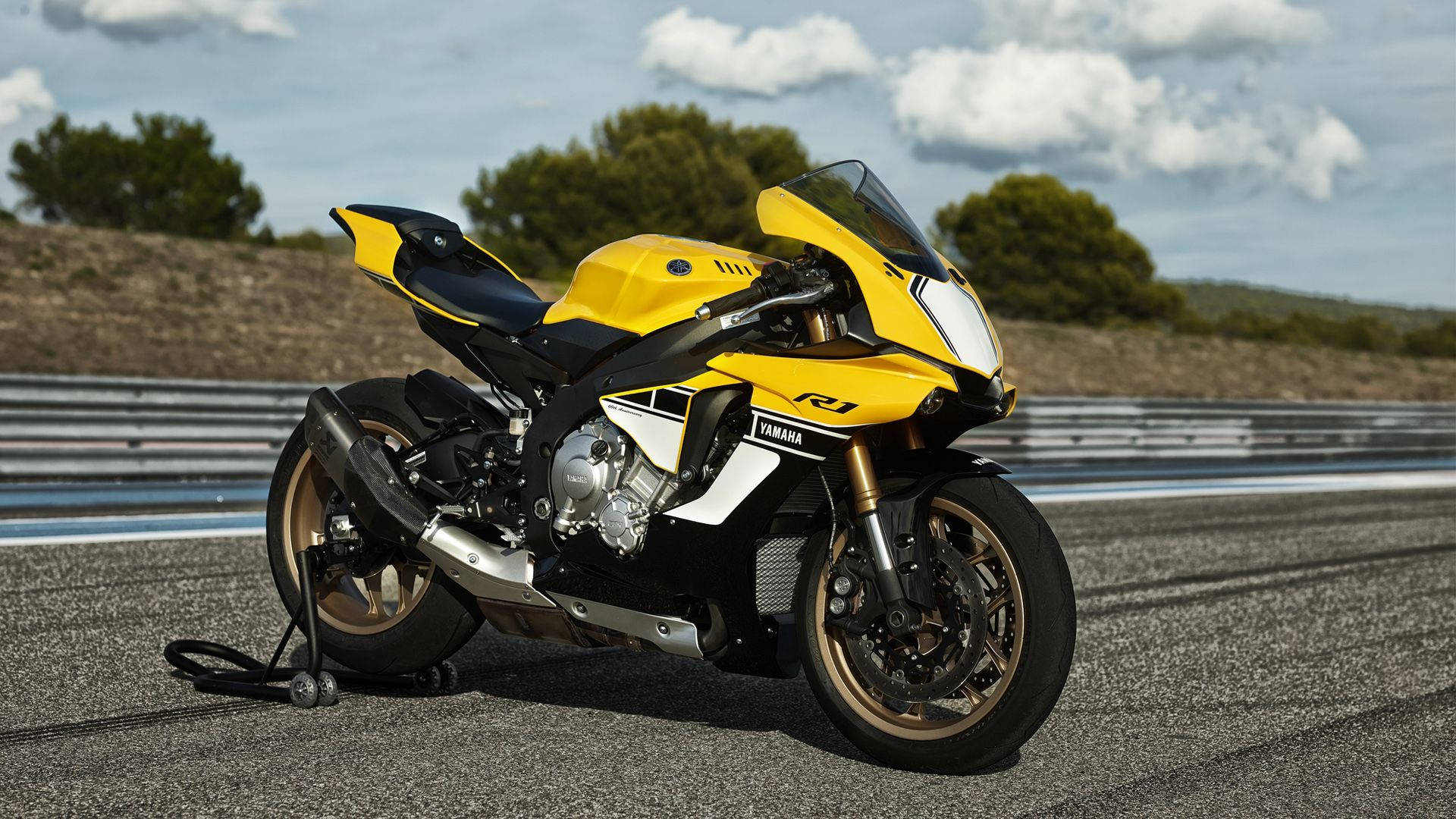 機車 桌布 壁紙 — 一輛鮮黃色的Yamaha R1摩托車停放在有護欄和綠色山丘的賽道上，天空局部多雲。