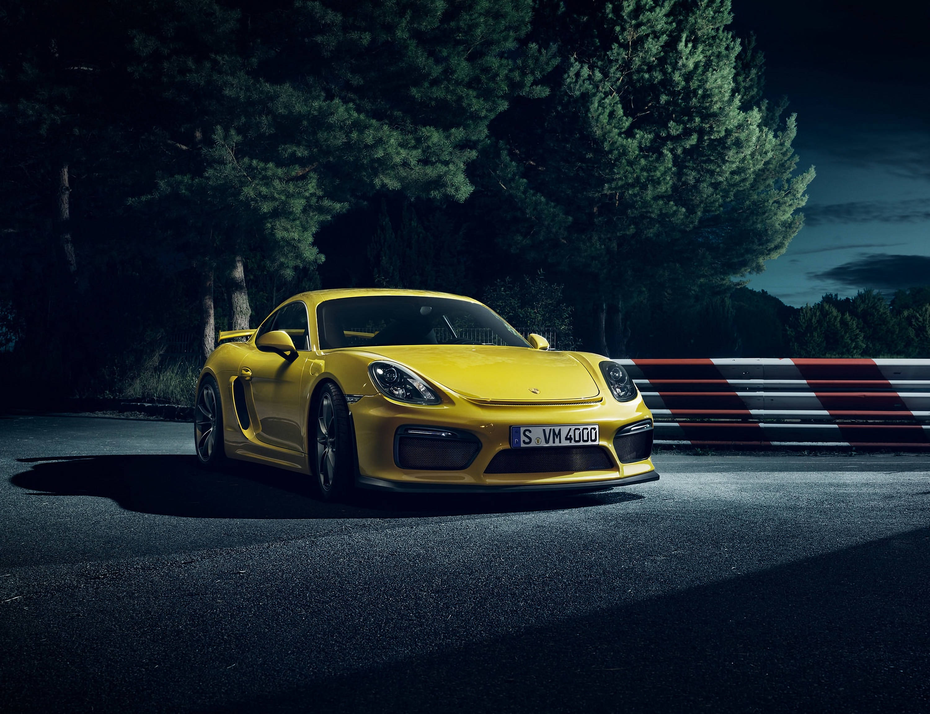 保時捷 桌布 壁紙 — 一輛鮮黃色的保時捷 Cayman GT4 停在夜間賽道上，周圍有深色樹木和紅白相間的護欄。