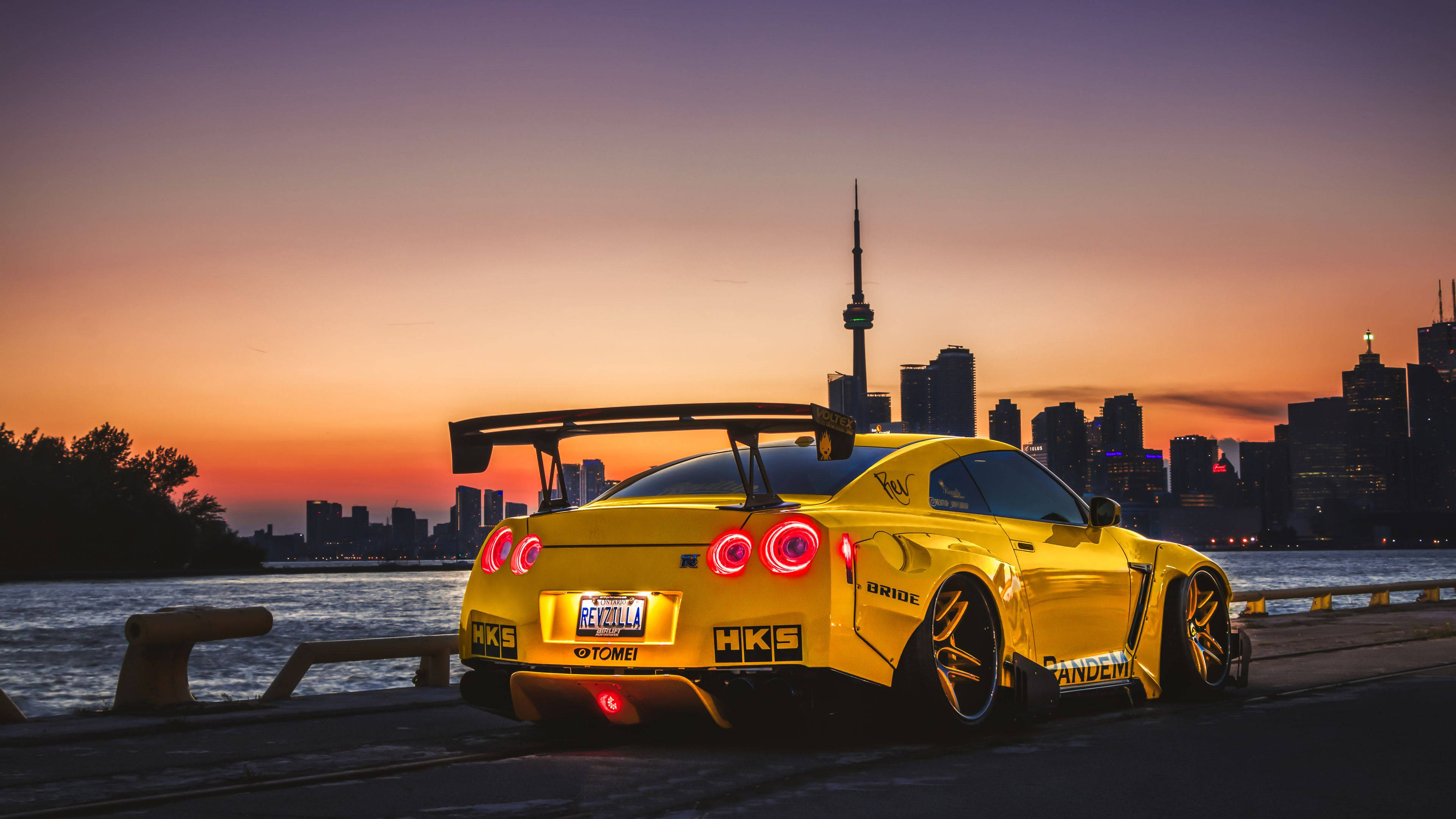 Nissan 桌布 壁紙 — 一輛鮮黃色的日產GT-R，亮起紅色尾燈，停泊在海濱長廊上，背景是多倫多天際線和CN塔，夕陽西下。