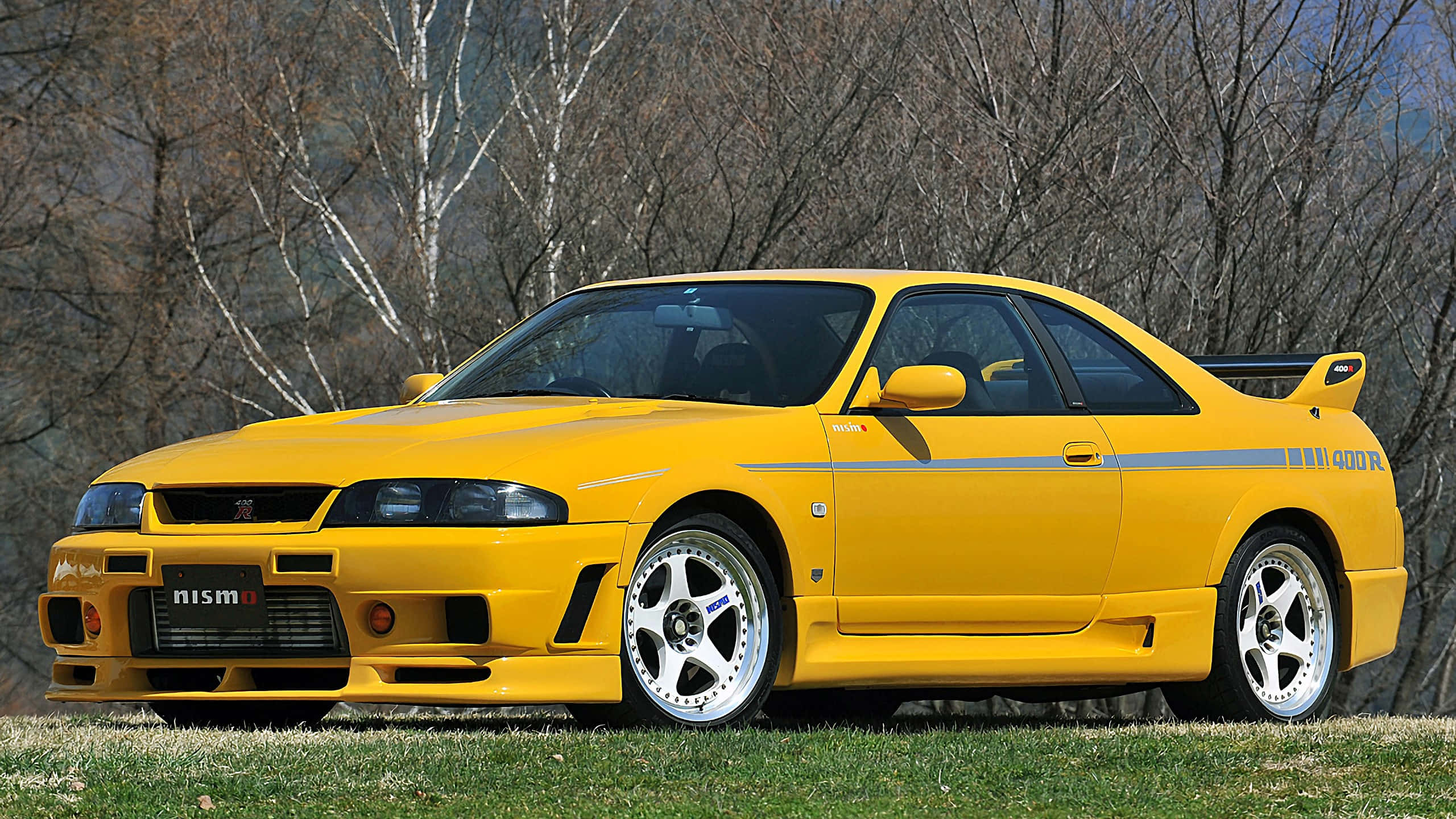 R33 GT-R 桌布 壁紙 — 一輛鮮黃色的日產Skyline R34 GT-R跑車停在草地上，背景有光禿禿的樹木。