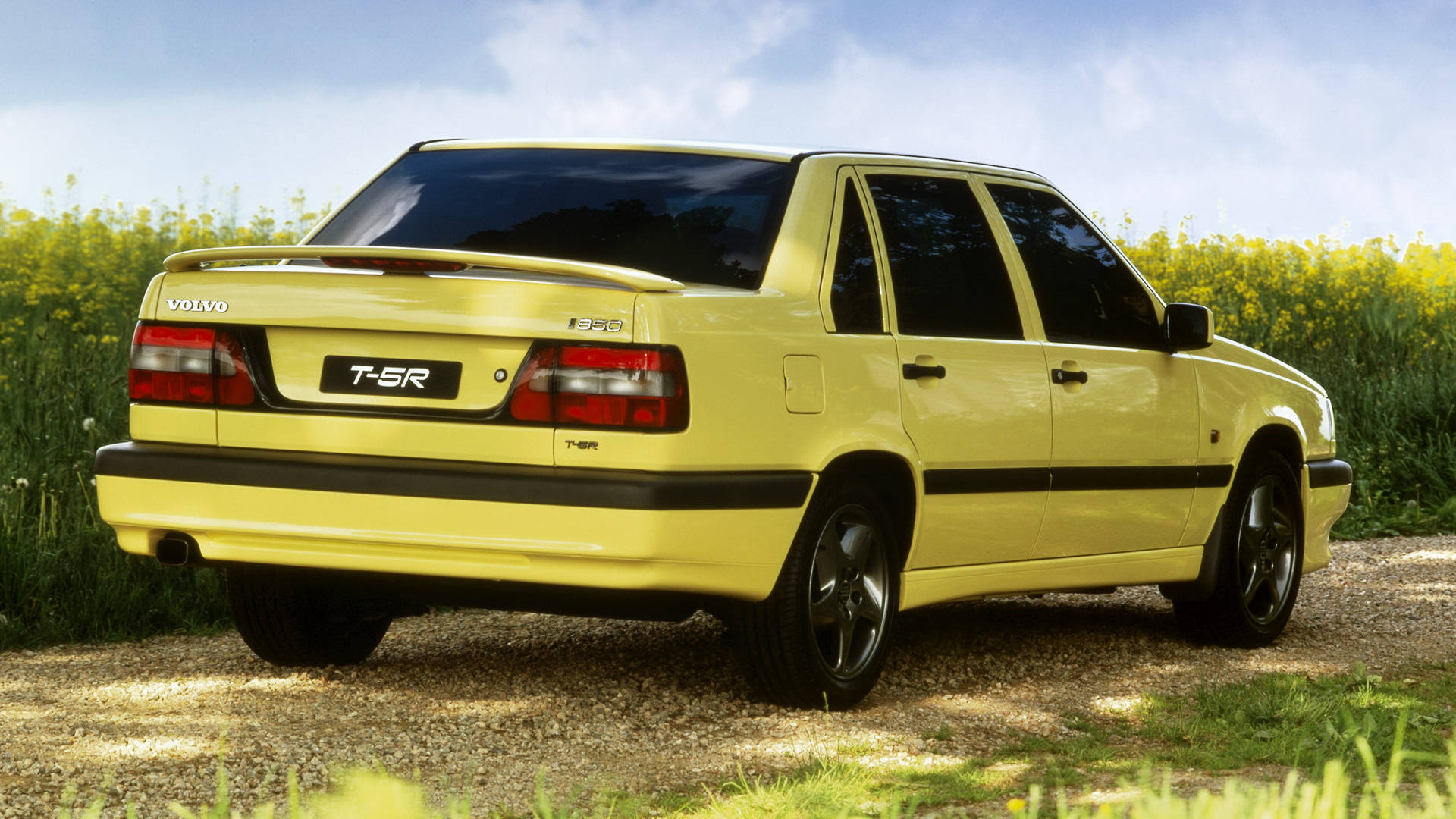 volvo iPhone 桌布 壁紙 — 一輛鮮黃色的沃爾沃 T5R 房車停泊在碎石路上，旁邊是盛開的黃色花田，上方是明亮的藍天。