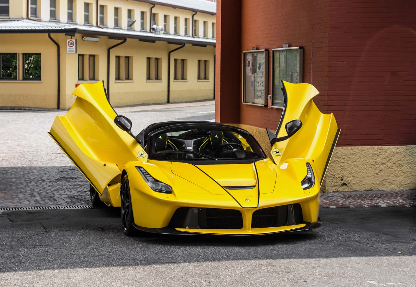 法拉利 桌布 壁紙 — 一輛鮮黃色的法拉利 LaFerrari 打開鷗翼車門，停在紅磚建築旁的鵝卵石街道上。