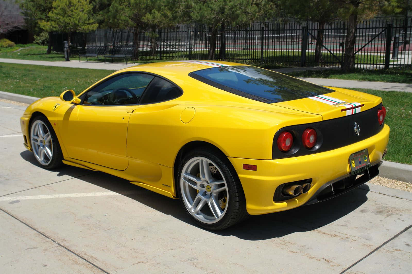 Ferrari 360 Modena 桌布 壁紙 — 一輛鮮黃色的法拉利360跑車停放在陽光明媚的混凝土車道上，背景有綠樹和圍欄。