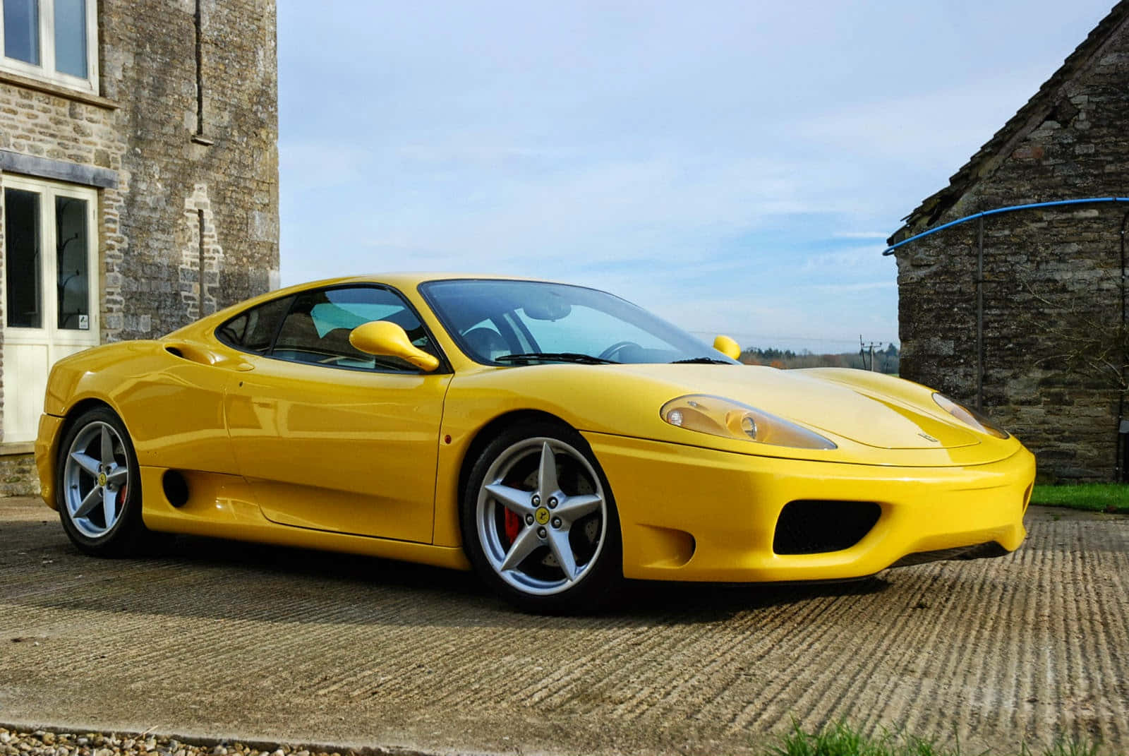 Ferrari 360 Modena 桌布 壁紙 — 一輛鮮黃色的法拉利360跑車在日光下閃閃發光，停泊在質感車道上，旁邊是質樸的石頭建築。