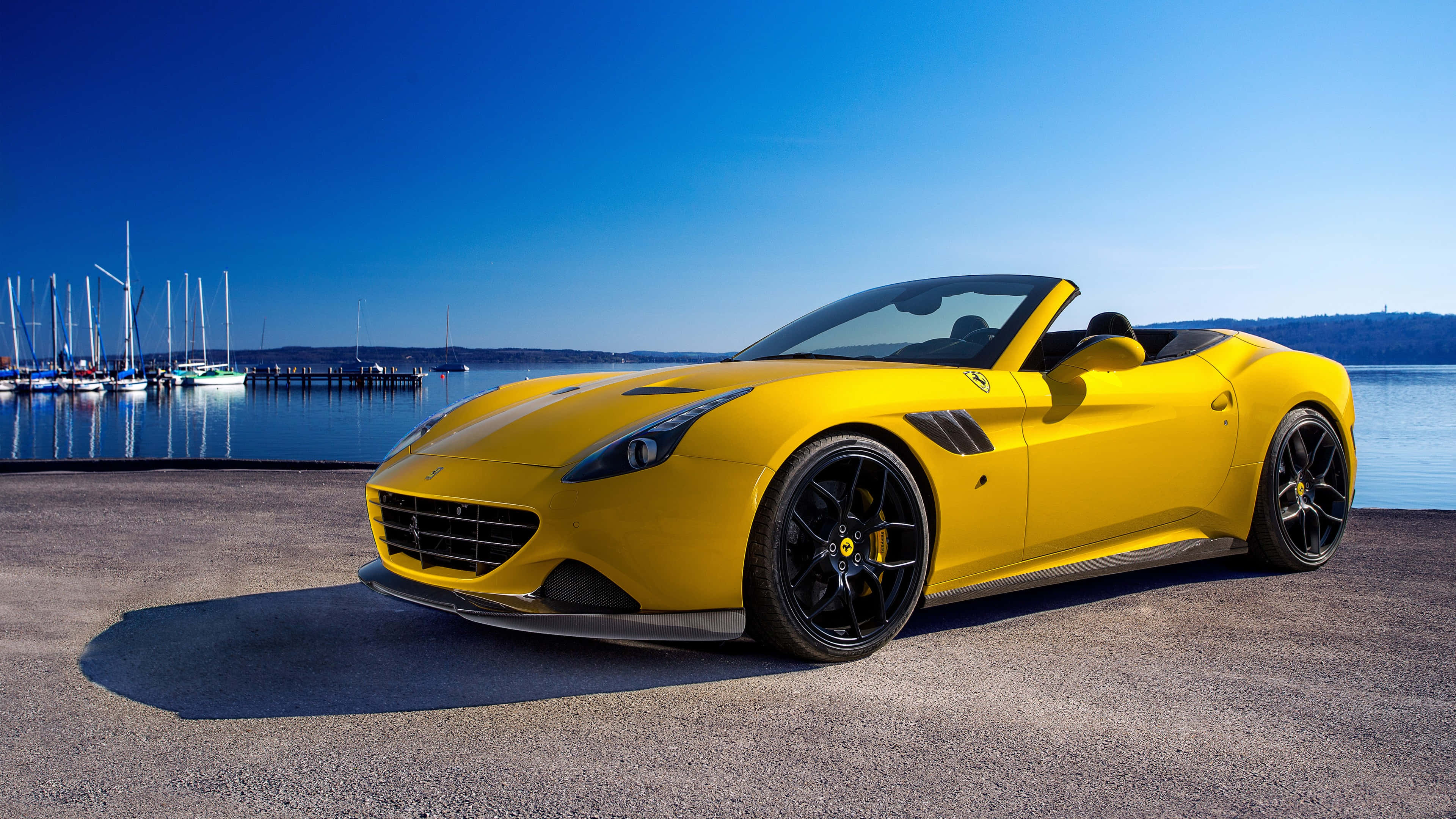 Ferrari Portofino 桌布 壁紙 — 一輛鮮黃色的法拉利敞篷車停泊在碼頭上，旁邊有帆船和寧靜的水面，天空清澈湛藍。